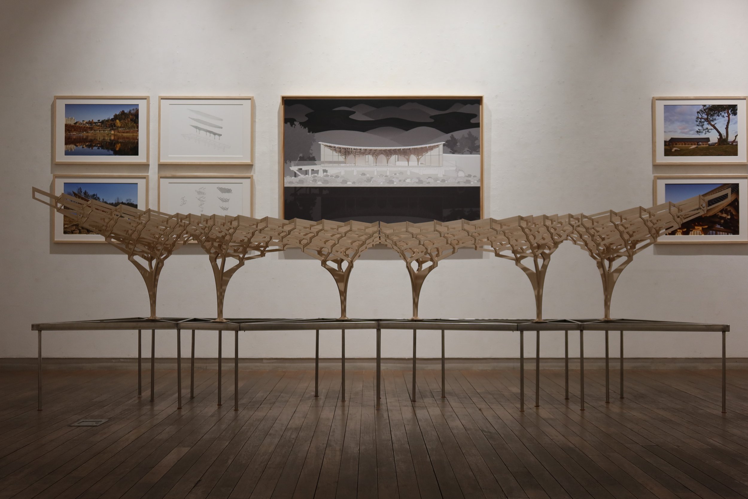 A gallery wall with six framed photos and a sculptures of a bridge on a table in front. The photos depict a landscape with water and trees, and architectural designs.