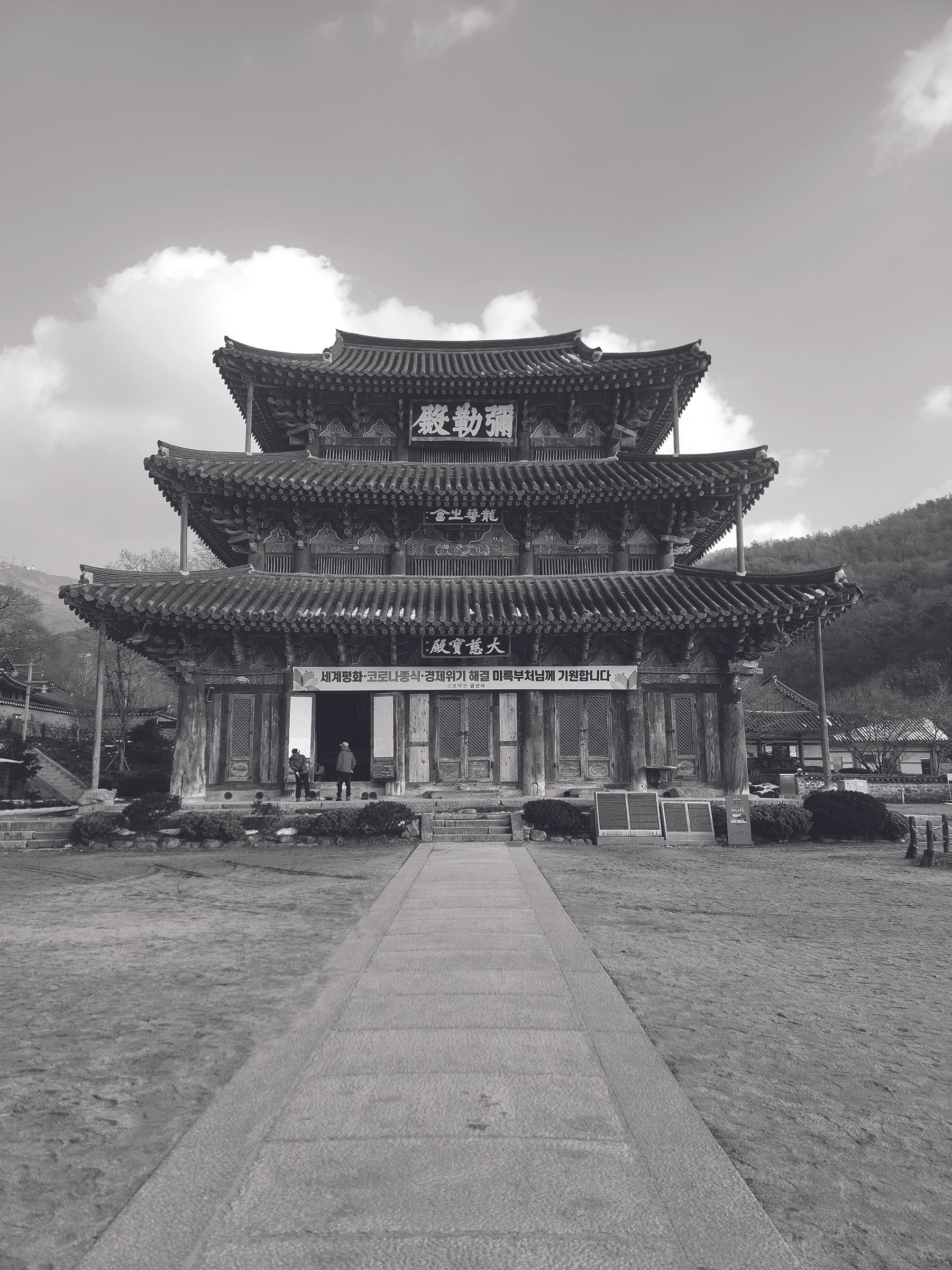 Black and white photo of a traditional East Asian pagoda-style building with multiple tiers, ornate roof eaves, and decorative details, set against a cloudy sky and distant hills.