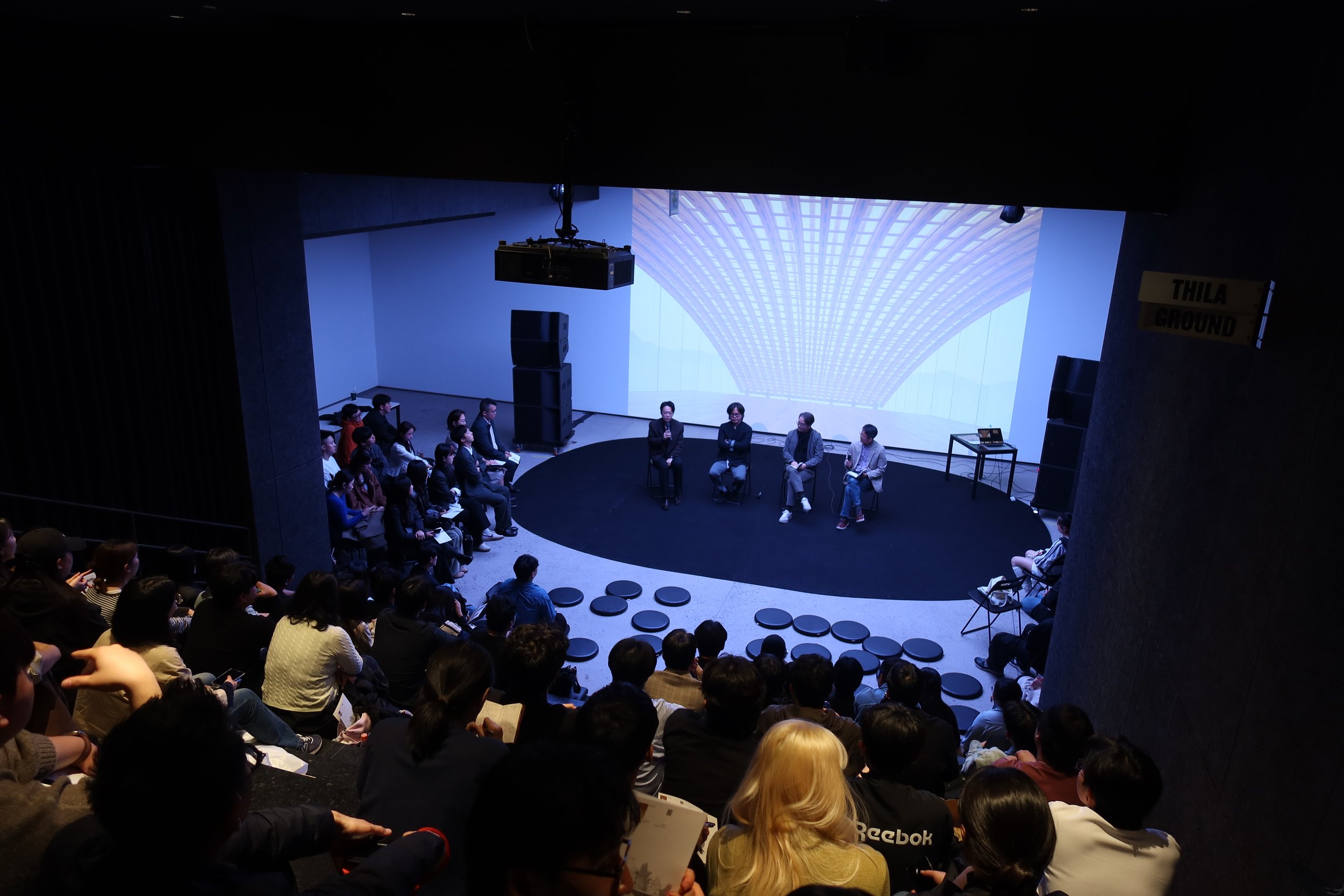 A large group of people attending an indoor event, seated and facing a stage where five individuals are engaged in a panel discussion. The background features a large screen displaying a digital architectural image.