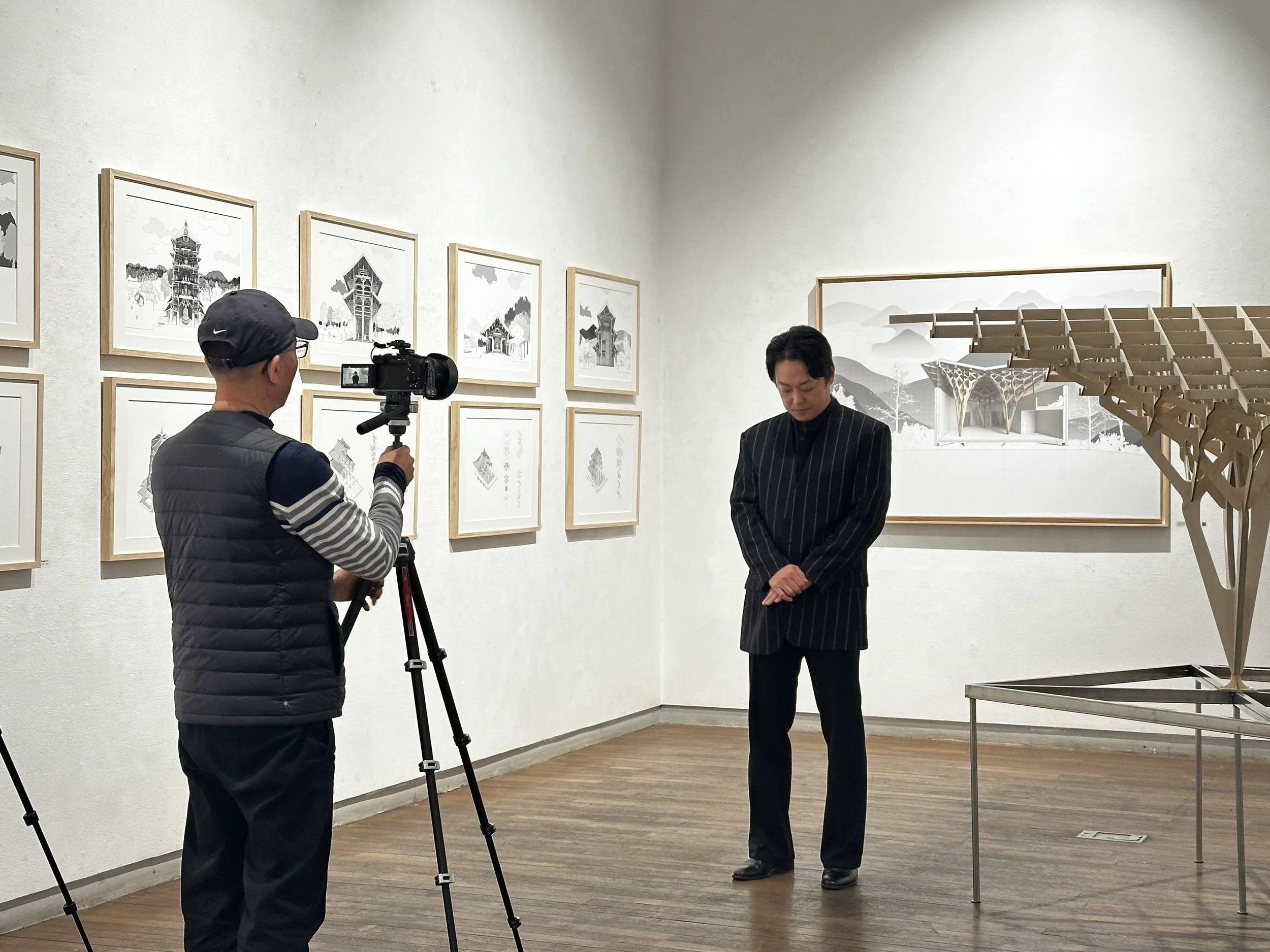 Two men in an art gallery, one recording with a camera and the other looking down, standing near framed artwork and a large wooden sculpture on display.