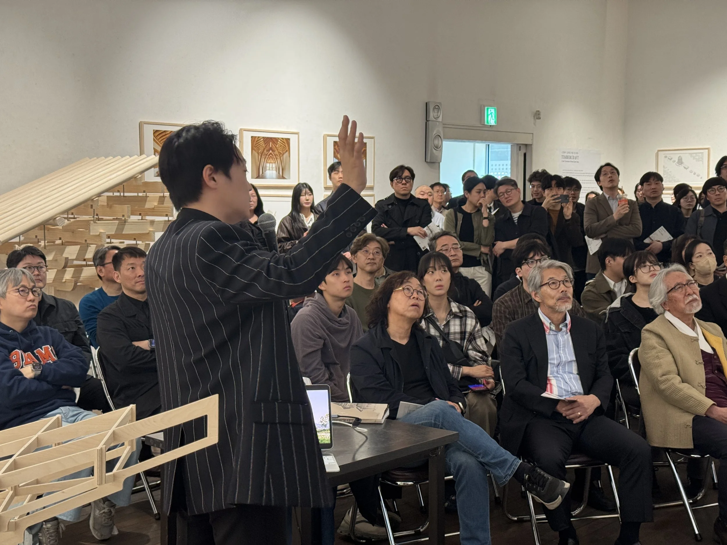 A person holding a microphone and raising their hand while speaking to a large group of seated and standing people in a room with framed pictures on the wall.