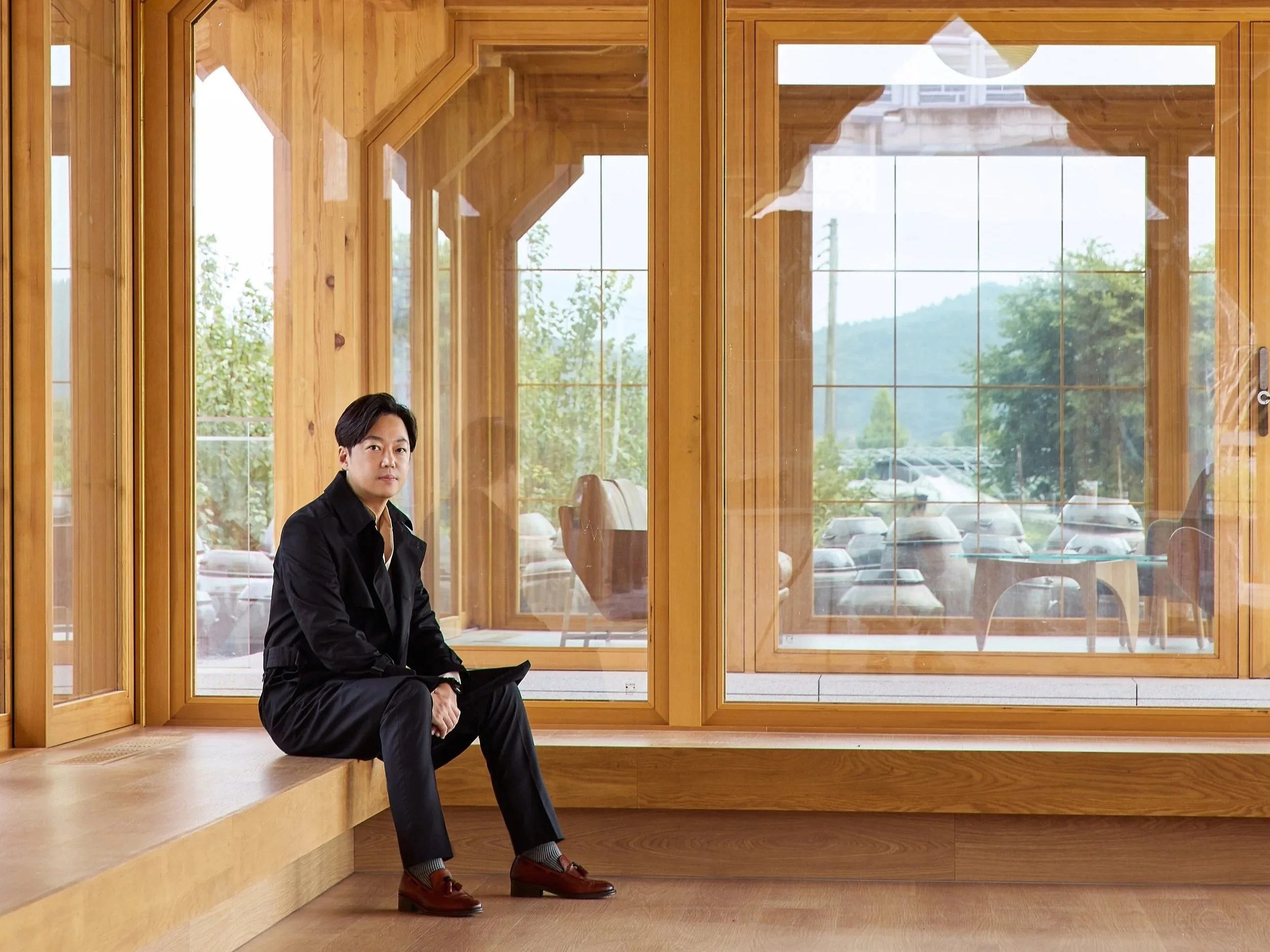A man with black hair sitting on a wooden bench inside a glass and wood structure, with trees and parked cars visible outside.