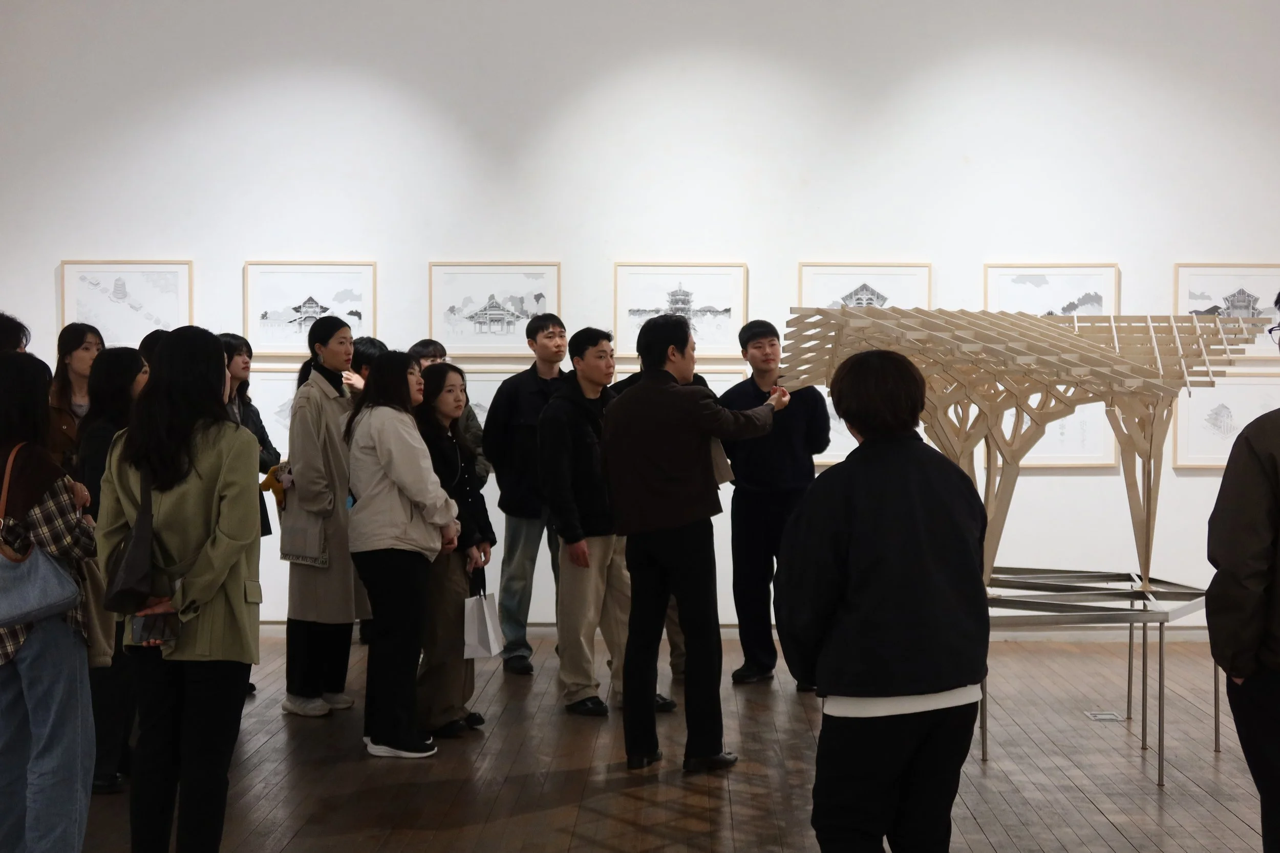 Group of people observing an art installation in a gallery, with framed pictures of traditional Asian pagodas on the wall behind them.