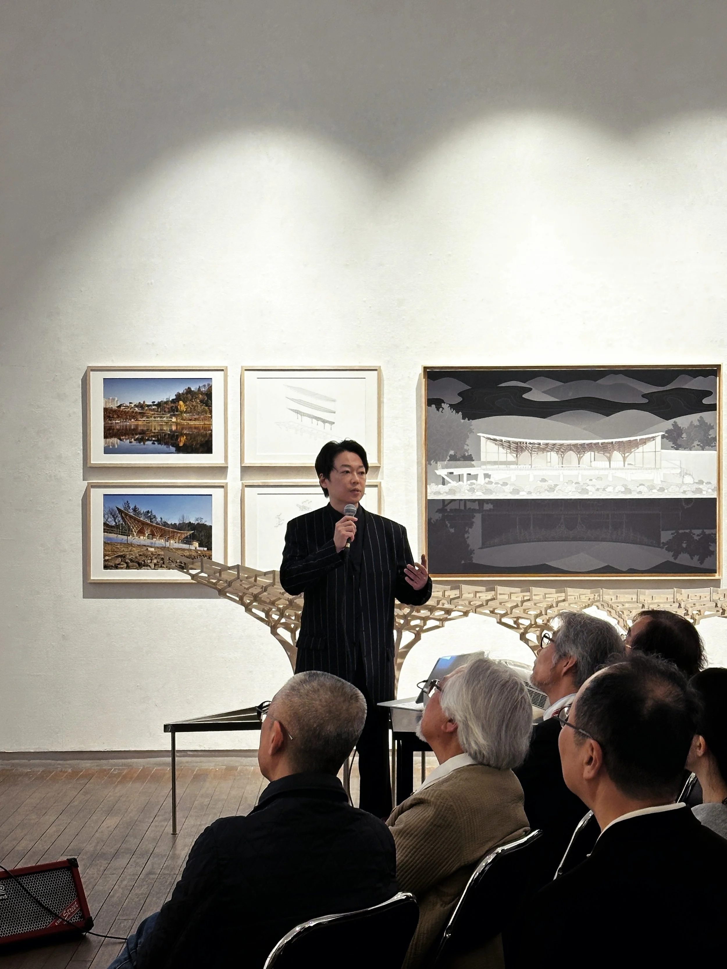A man giving a presentation at an art gallery or museum. He is holding a microphone and standing in front of framed artwork on the wall. Audience members are seated and listening.
