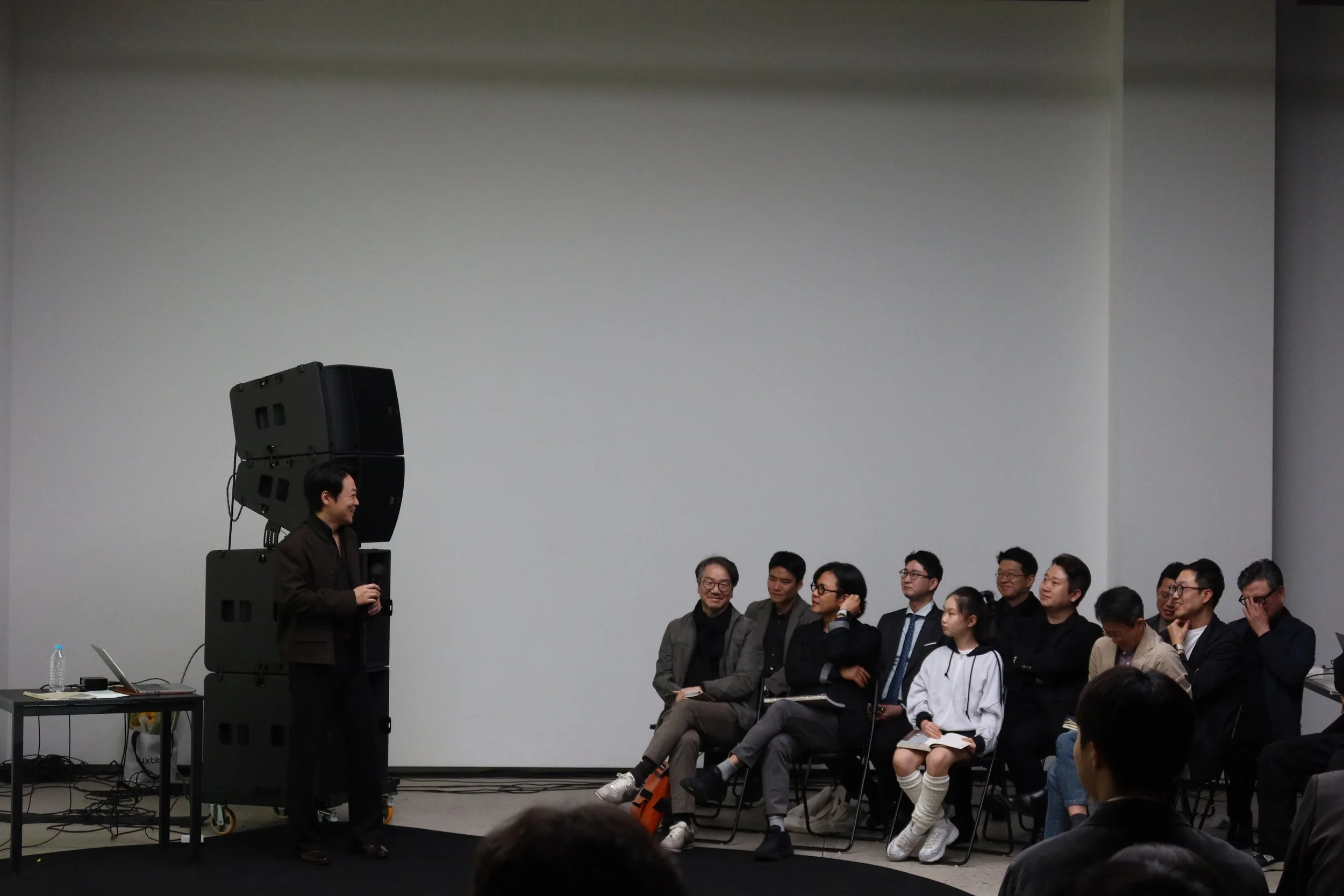 A group of people sitting and standing in a room, listening to a woman speaking at an event or presentation. The woman is standing near some large speakers and is holding a microphone, with a large blank wall behind her.