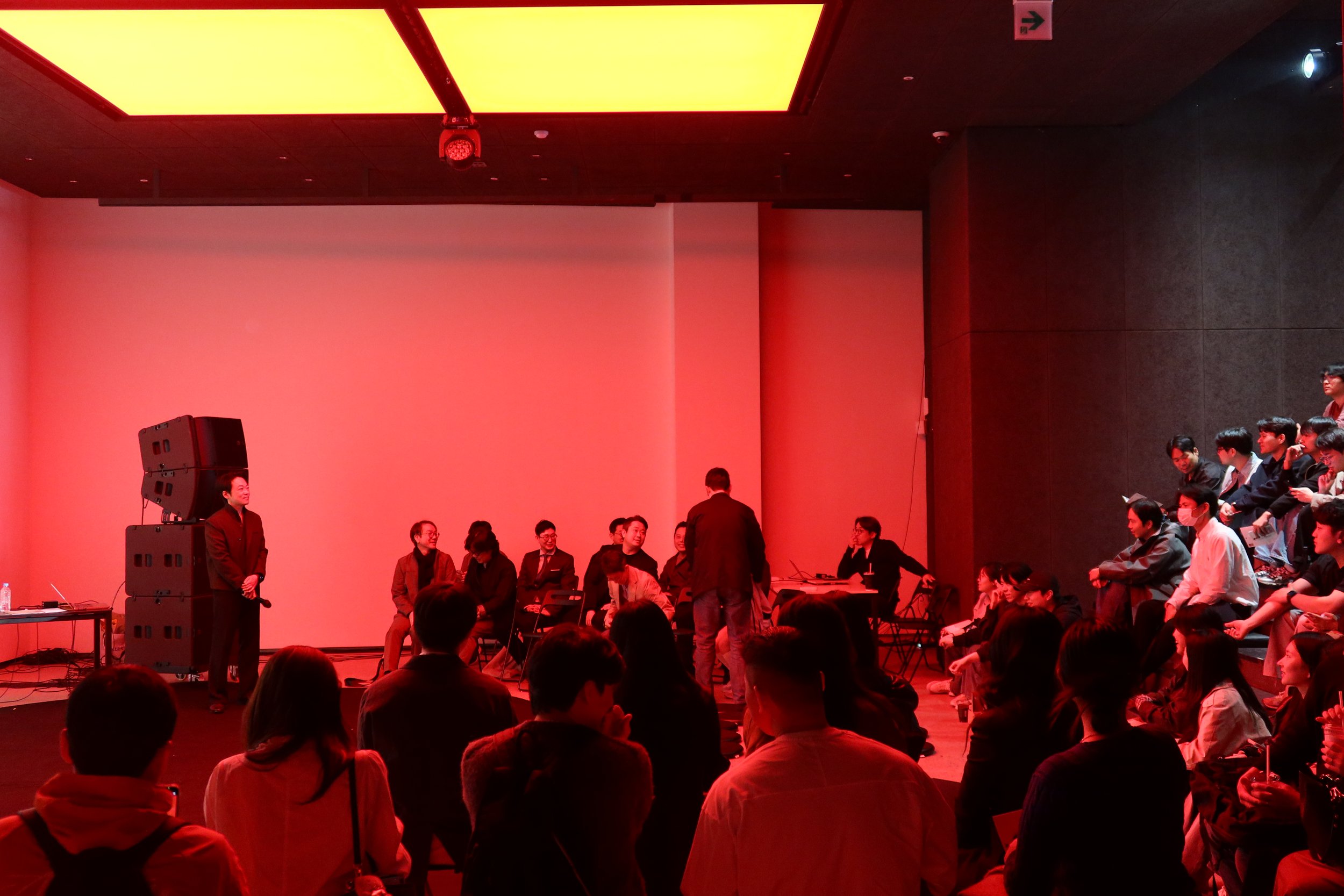 A group of people gathered in a room with red and yellow lighting. Some people are seated on chairs, while others are standing and appear to be engaged in conversation. The room has a dark wall on the right and a large yellow light panel on the ceili