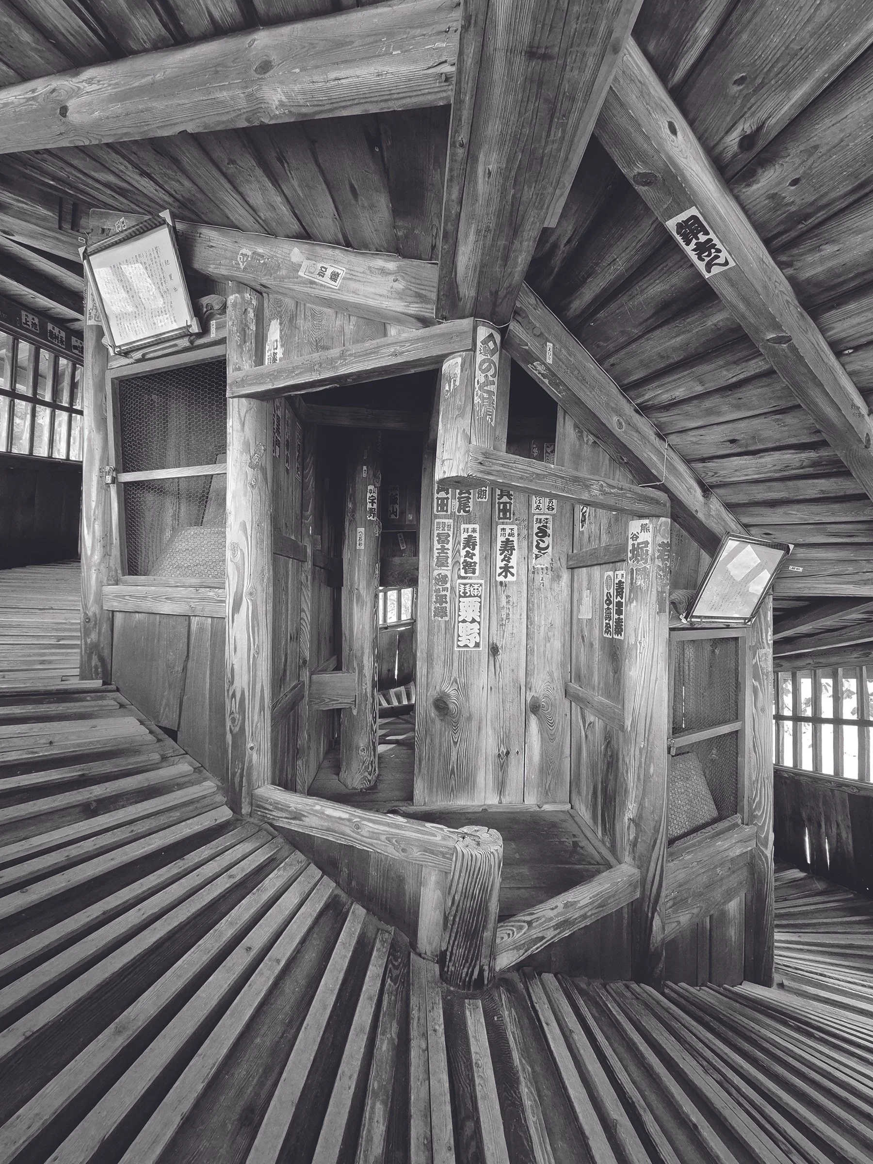 Black and white photo showing the interior of a traditional Japanese wooden structure with wooden beams, signs with Japanese writing, and a sliding window.