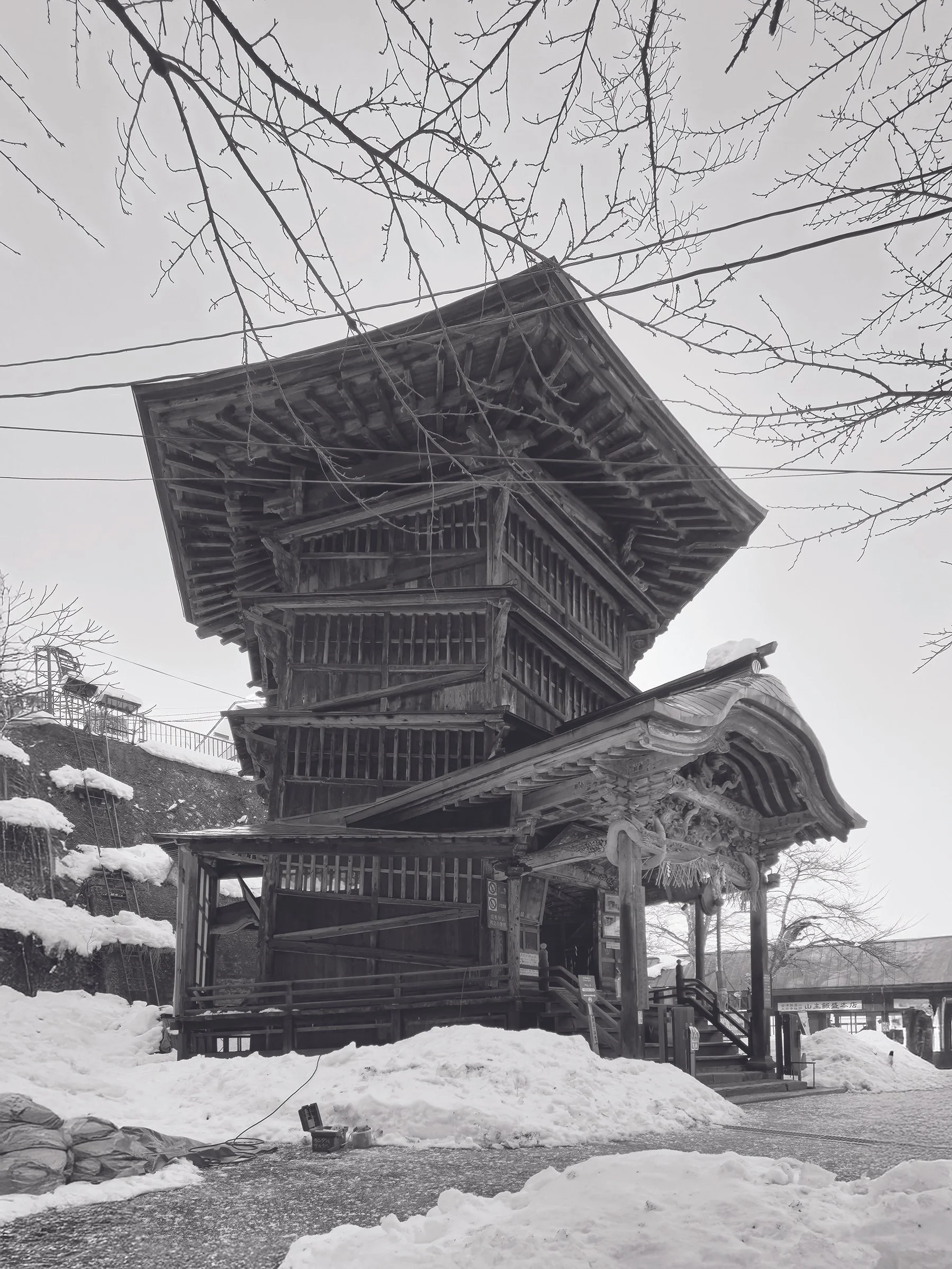 Traditional Japanese wooden temple or shrine structure with multiple tiers, surrounded by snow, with leafless trees and electrical wires in a winter setting.