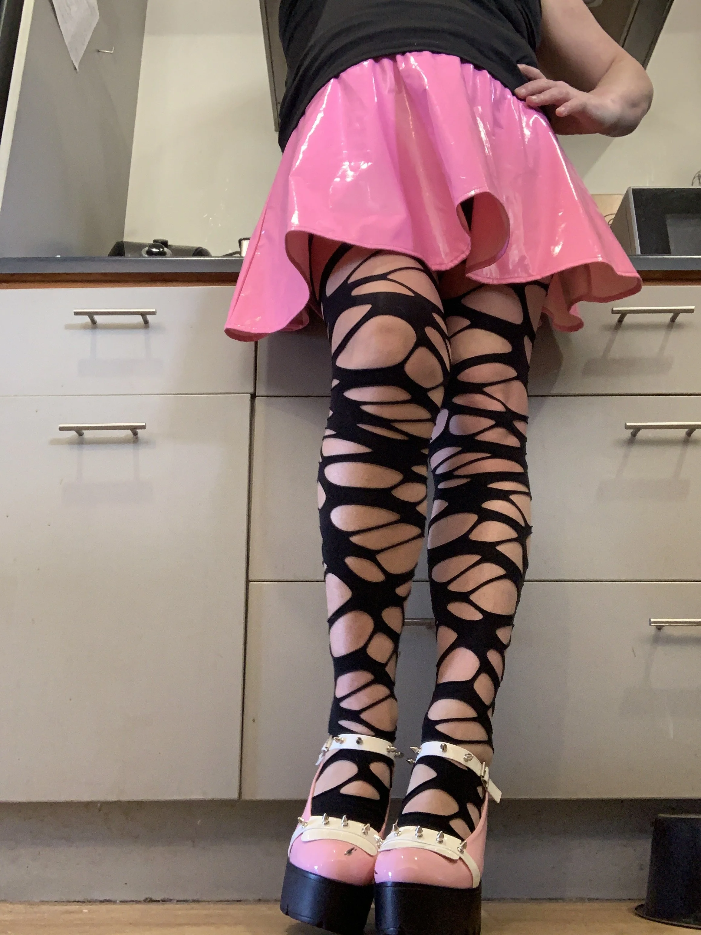 Woman standing in a kitchen wearing a pink shiny skirt, black fishnet tights with large holes, and pink platform shoes with black heels and white straps with silver spikes.