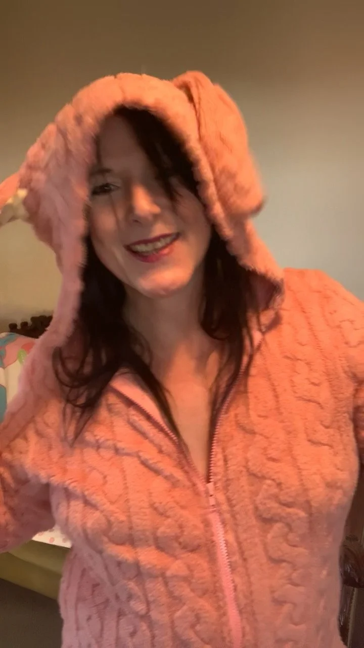 A woman smiling, wearing a pink, plush, hooded zip-up onesie, smiling