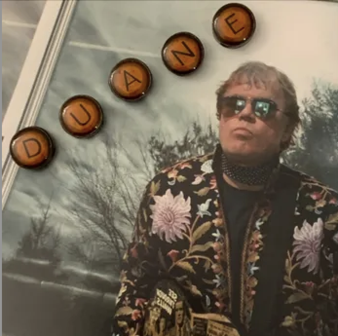 A person with sunglasses wearing a floral jacket standing next to a window reflecting trees. The top of the image shows four orange circular objects with black letters spelling 'D', 'U', 'A', 'N', 'E'.