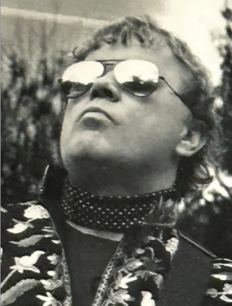 A person wearing sunglasses and a studded choker, in a confident pose.
