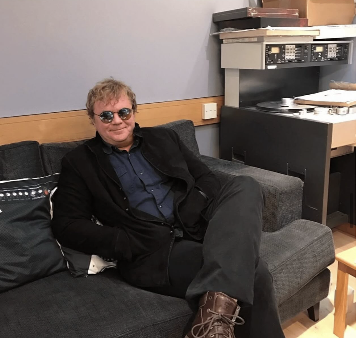 A man with curly blond hair and sunglasses sitting on a dark gray couch in a recording studio or radio station, with a vintage reel-to-reel tape recorder on a table behind him.