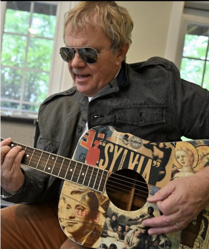 A man with blonde hair and sunglasses plays a decorated acoustic guitar in a room with windows.