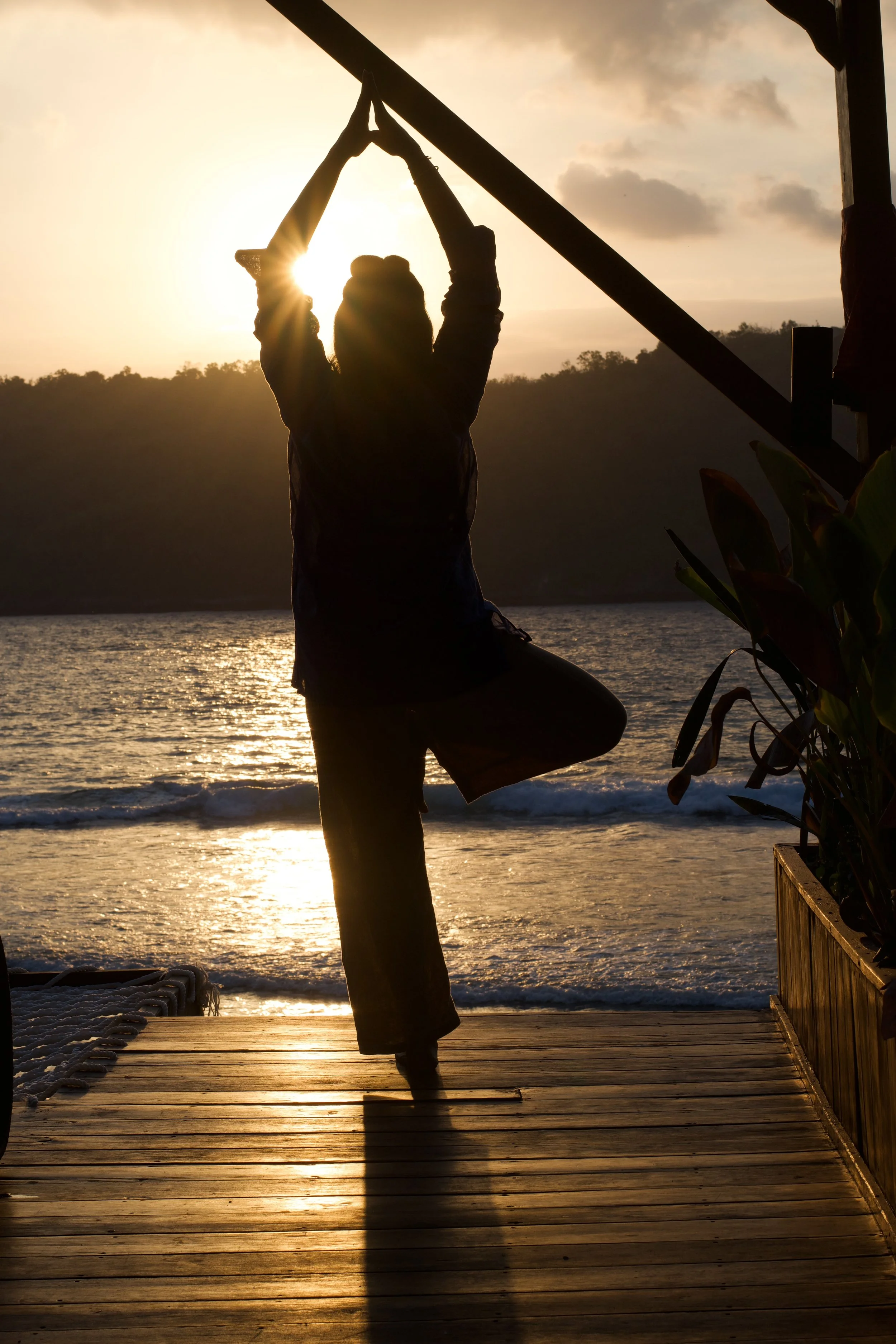 Silhouette of a person practicing yoga on a wooden deck at sunset with a body of water and hills in the background.