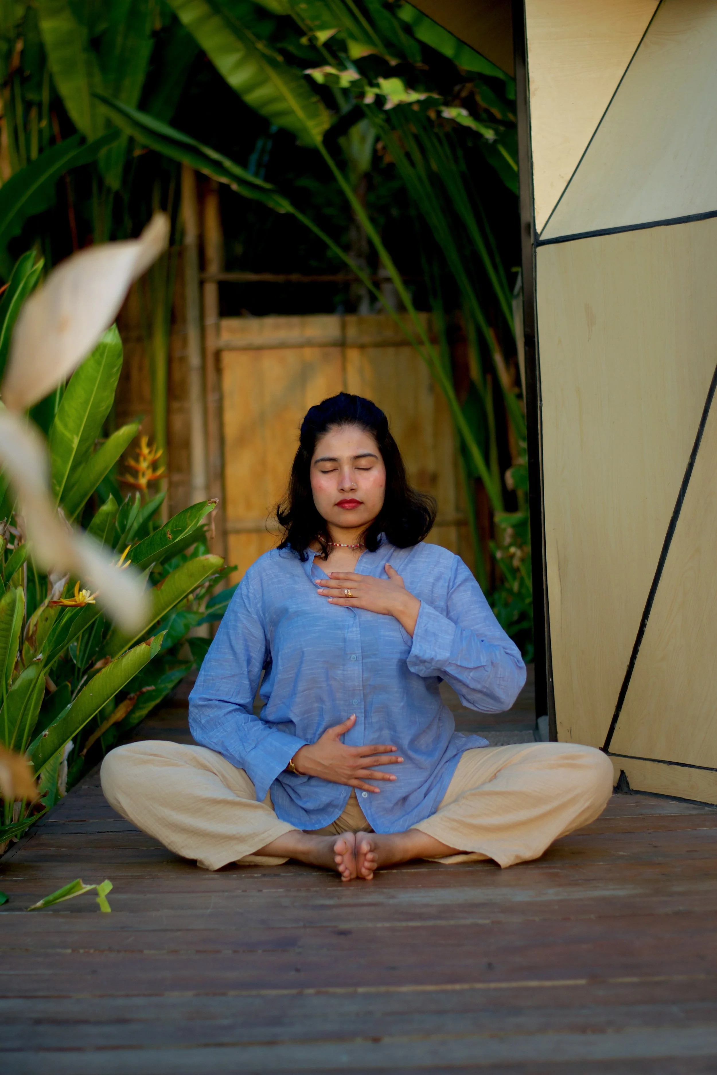 Woman meditating outdoors, sitting cross-legged on a wooden surface with her hands on her chest and stomach, surrounded by green foliage and plants.