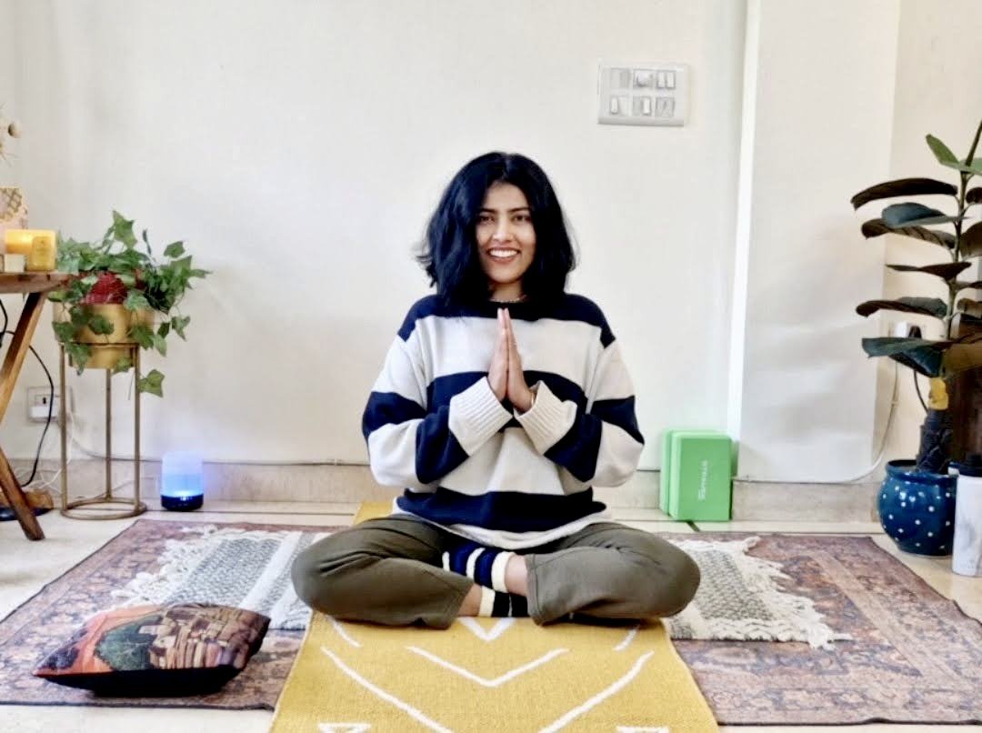 A woman sitting cross-legged on a yellow rug, practicing yoga or meditation with her hands in a prayer pose. She has shoulder-length dark hair and is smiling. The room has indoor plants, a small table, and a white wall.