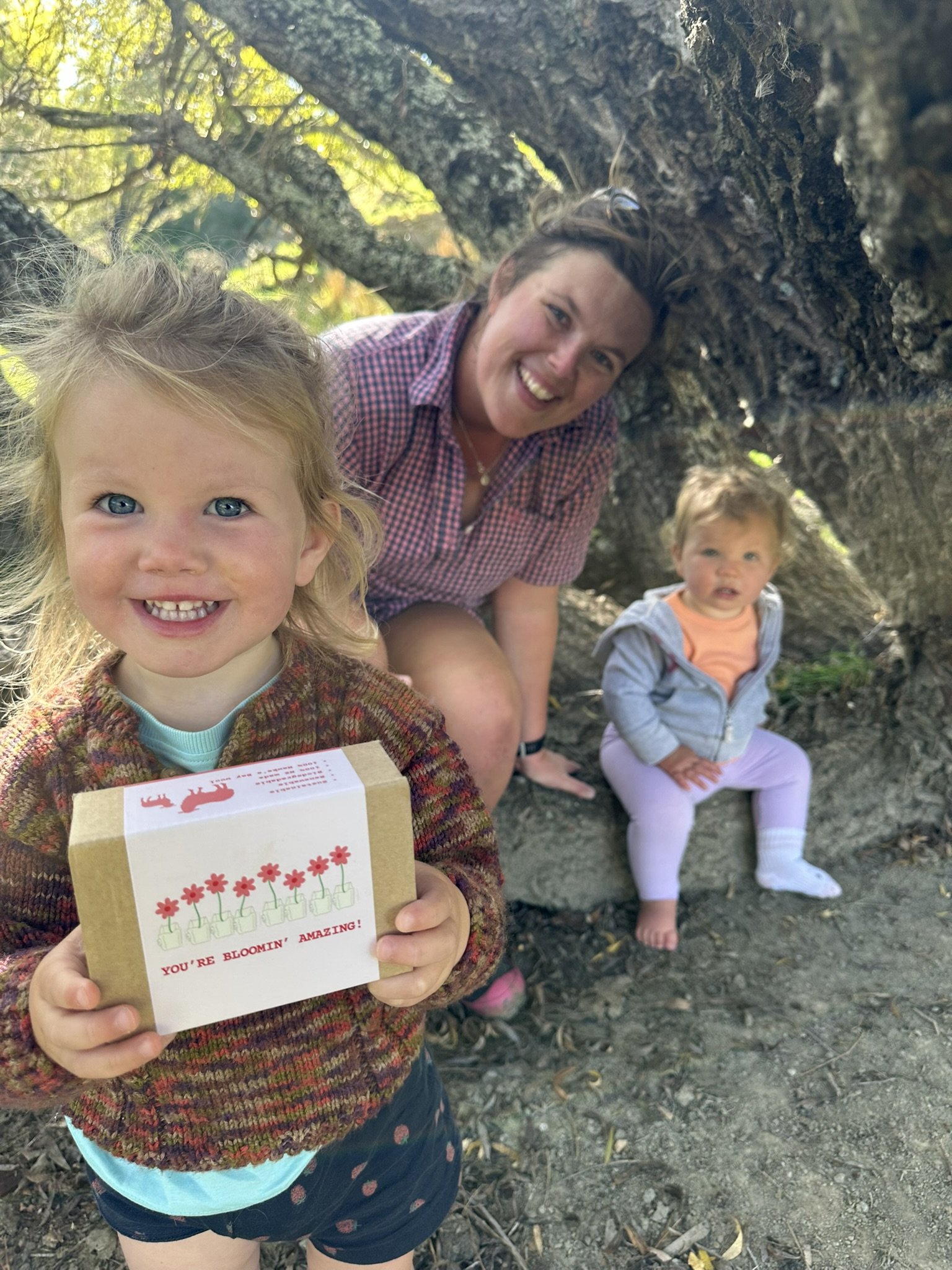 A young girl holding a birthday card with a birthday message, smiling at the camera. Behind her, an adult woman and another young girl are sitting under a large tree on a wooded ground with sunlight filtering through the leaves.