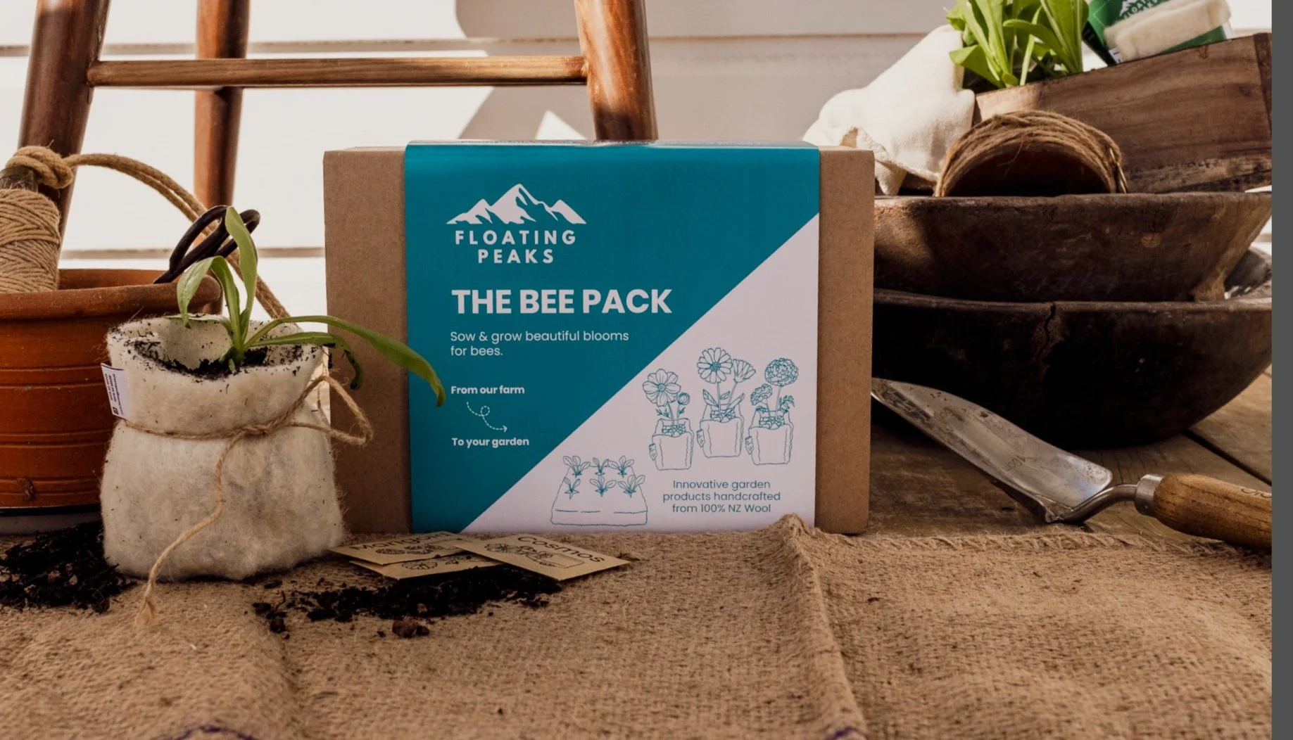 A cardboard box labeled "Floating Peaks, The Bee Pack" with illustrations of blooming flowers in pots, surrounded by gardening tools, a small potted plant, and wooden bowls on a rustic wooden surface.