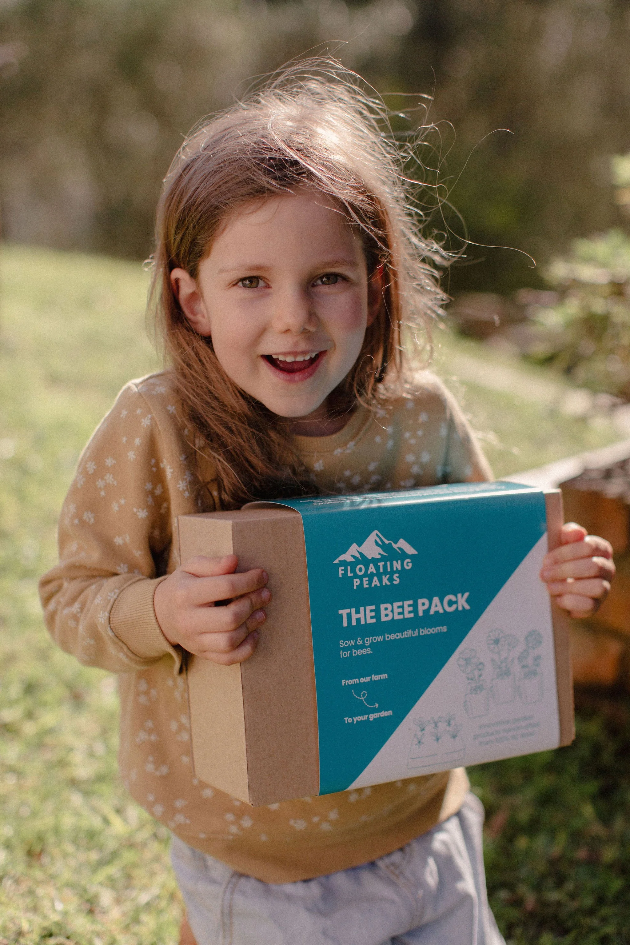 A young girl with long red hair holding a box labeled 'The Bee Pack' from Floating Peaks, outdoors on grass with sunlight.