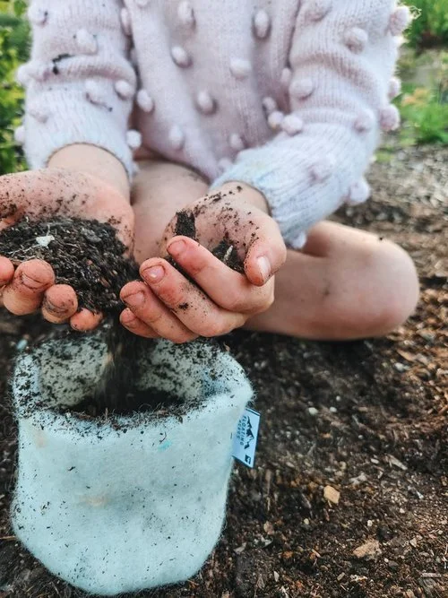 A child in a pink sweater with textured pom-pom details holds soil in both hands, pouring it into a white fabric pot outdoors.