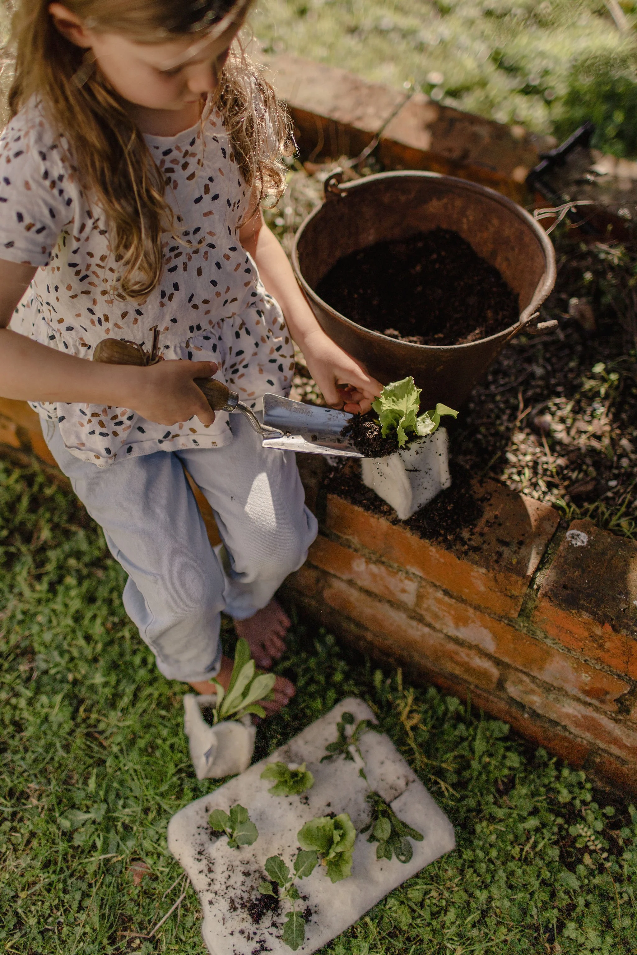 A young girl with glasses planting seedlings in a garden, with gardening tools, soil, and plants visible around her.