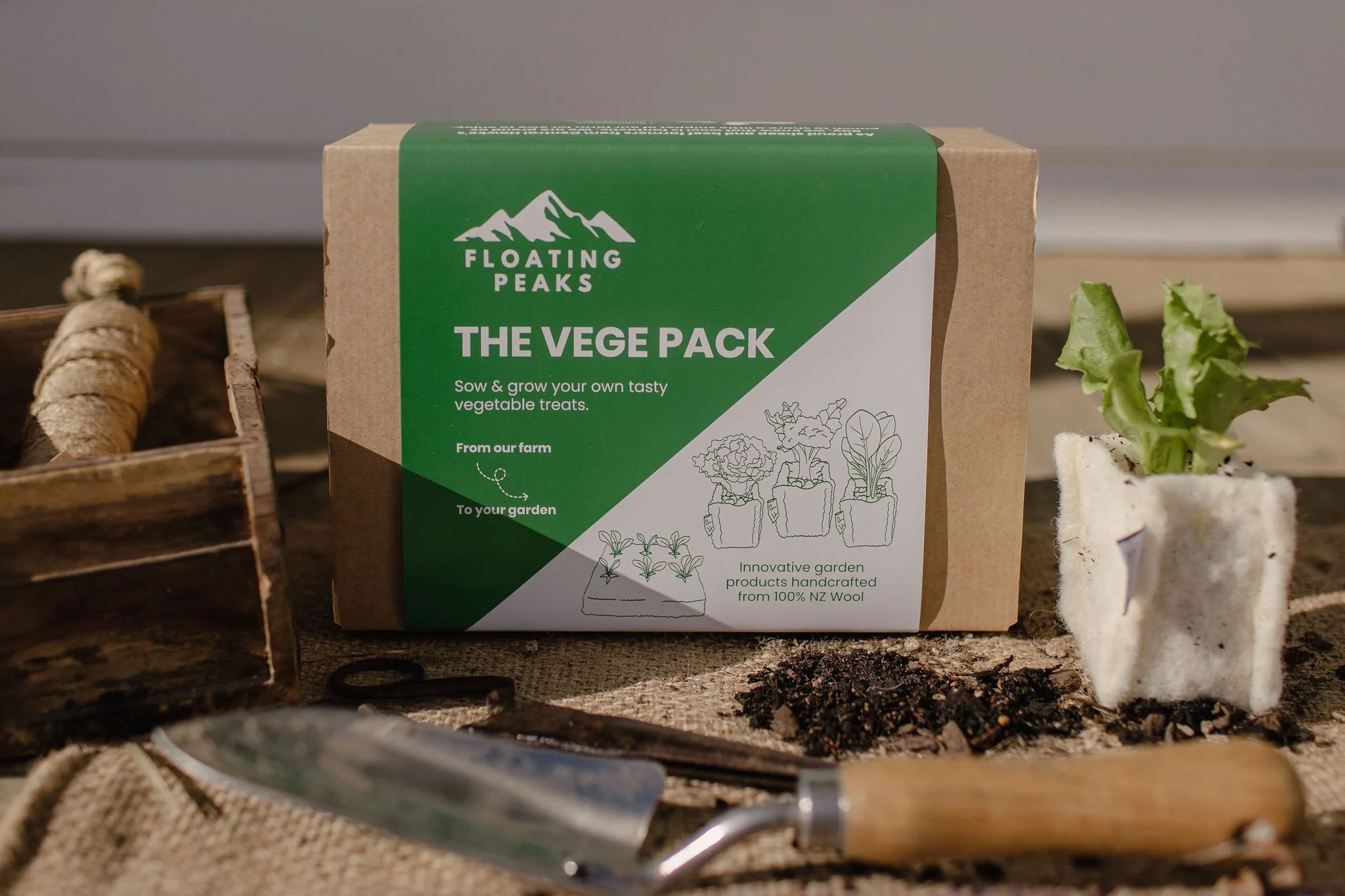 A cardboard packet of Floating Peaks 'The Veggie Pack' vegetable growing kit on a wooden surface next to a small plant in a fabric pot, gardening tools, and a wooden crate, with soil and gardening materials around.