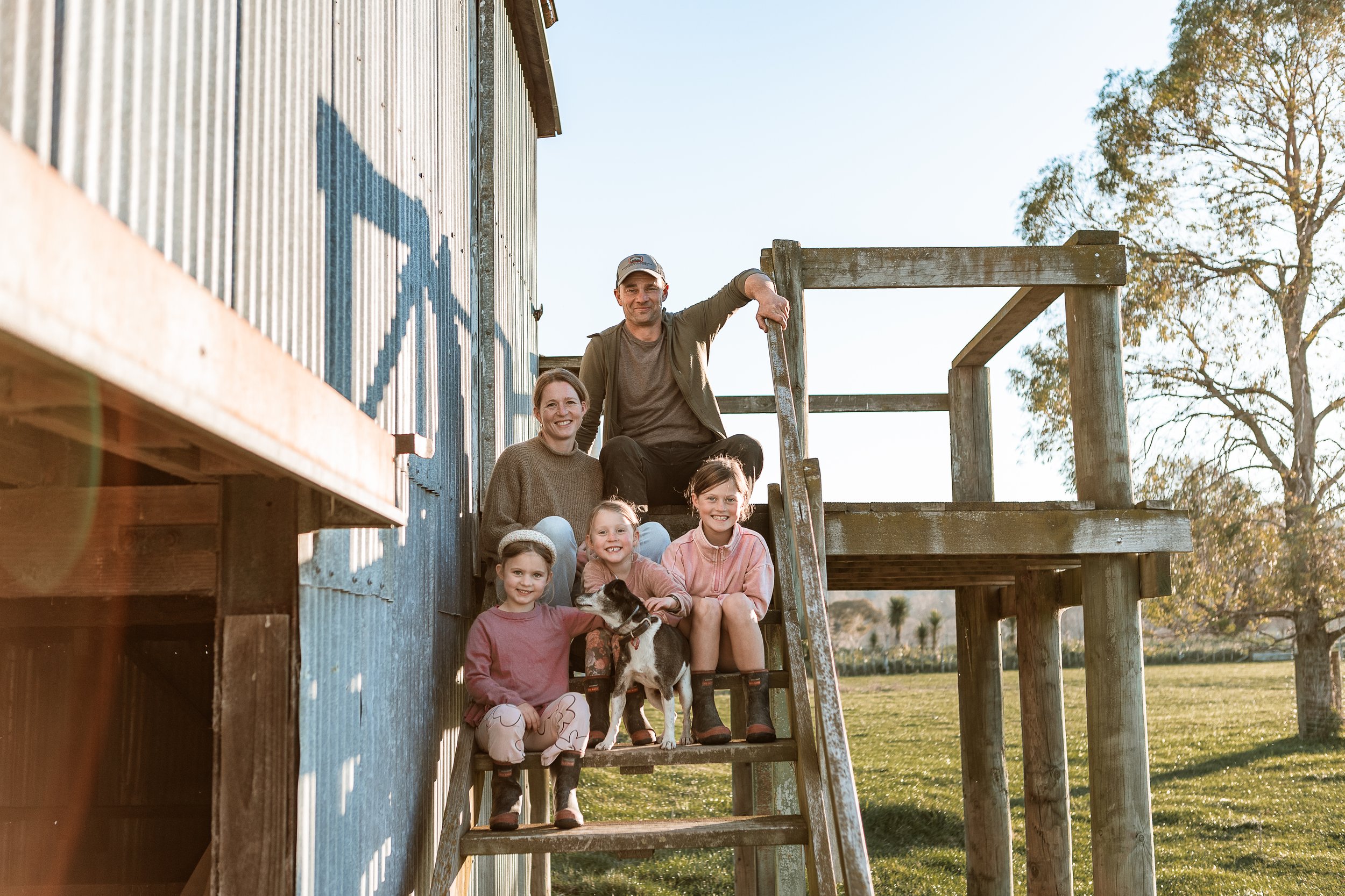 A group of children and adults sitting on wooden stairs outside a rustic building, with trees and a clear sky in the background.