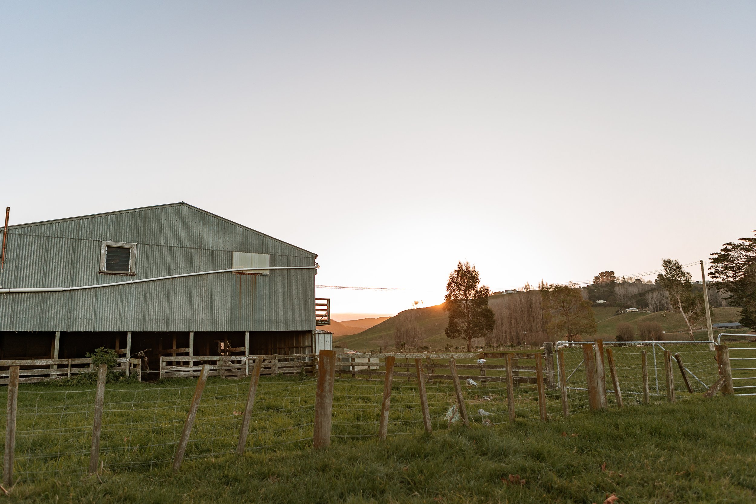 A rural scene at sunset featuring a large metal barn, rolling green hills, trees, and a wooden fence in the foreground.