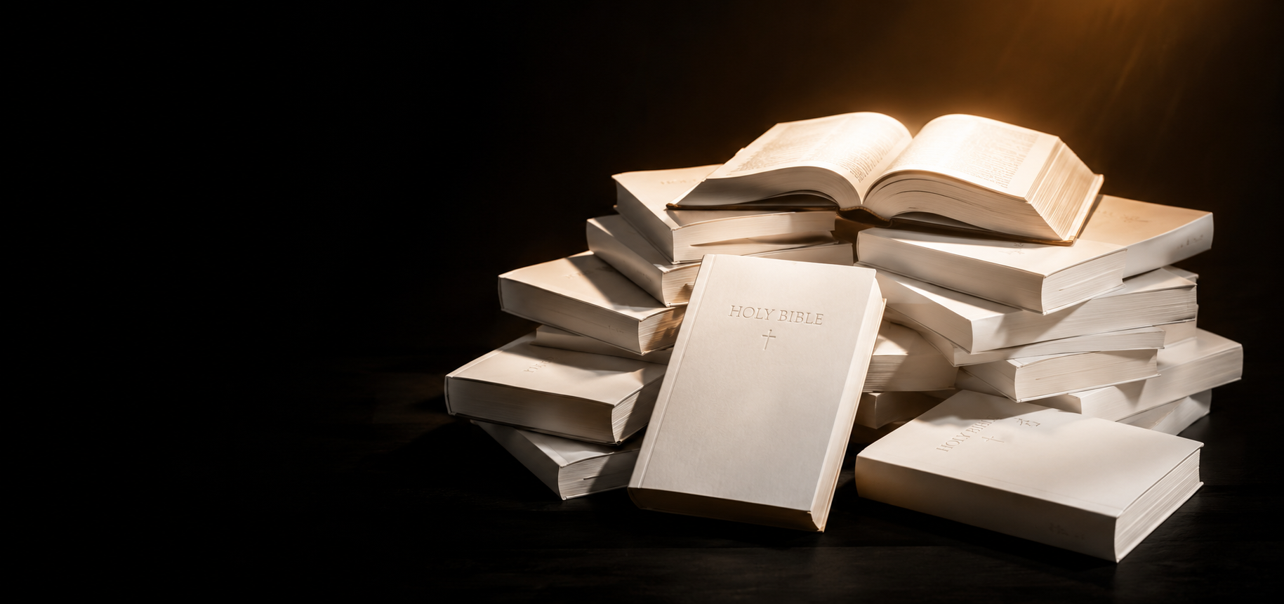 Stack of white books with one open Bible on top, illuminated by a warm light, against a dark background.