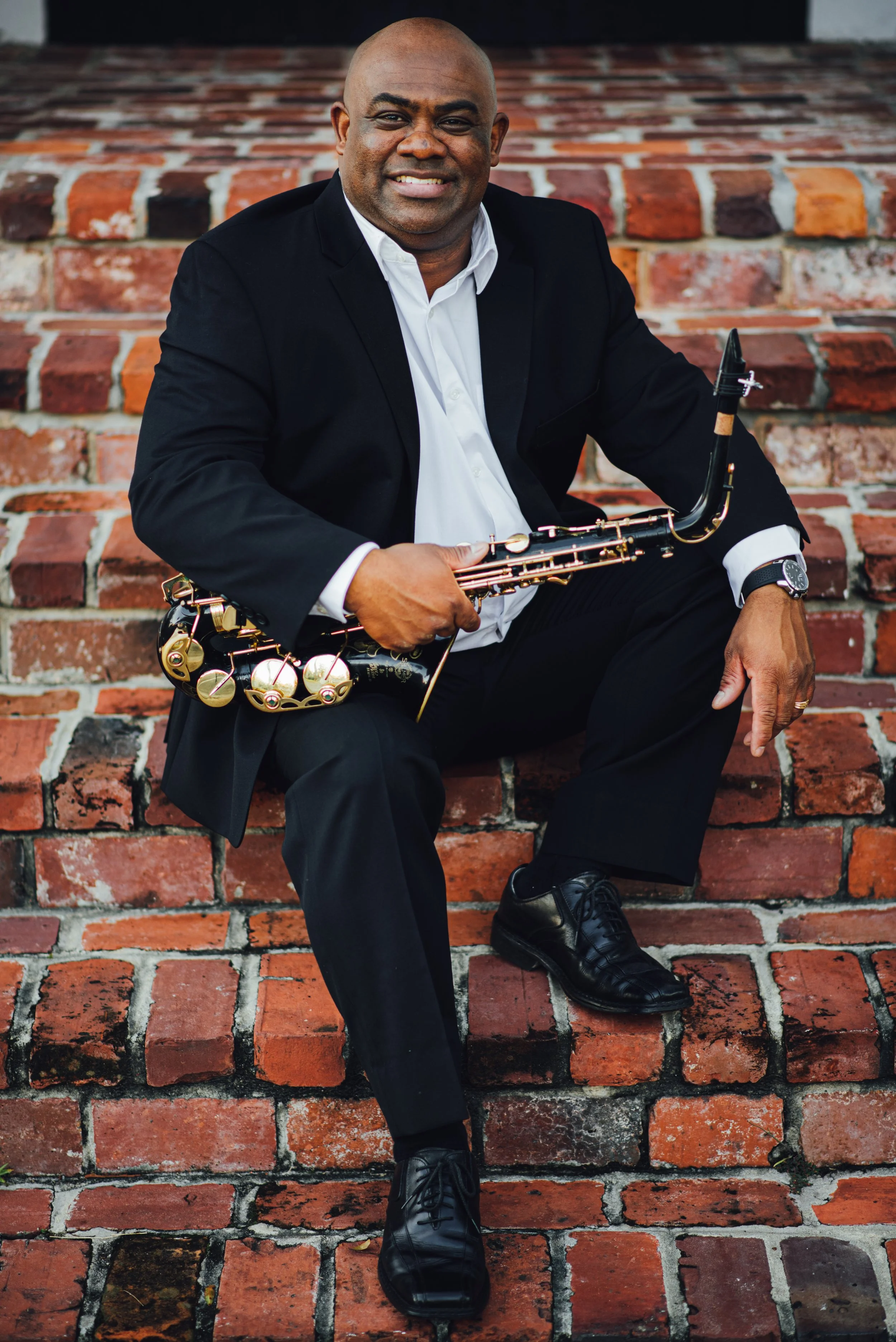 A man sitting on brick steps, holding a saxophone, wearing a black suit and white shirt, with a watch on his left wrist and smiling.
