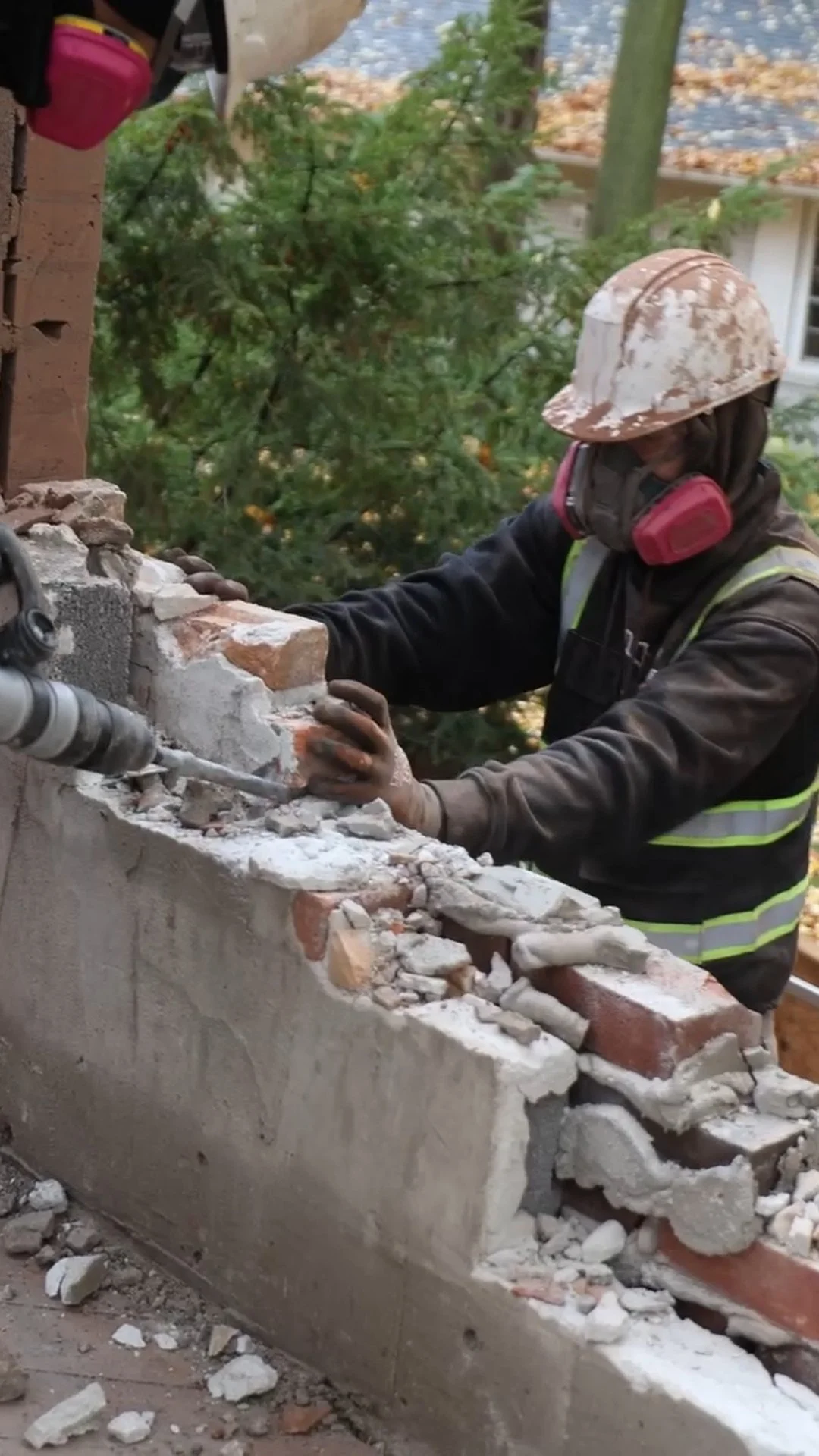 Worker in safety gear demolishing a brick and concrete wall with a power chisel.