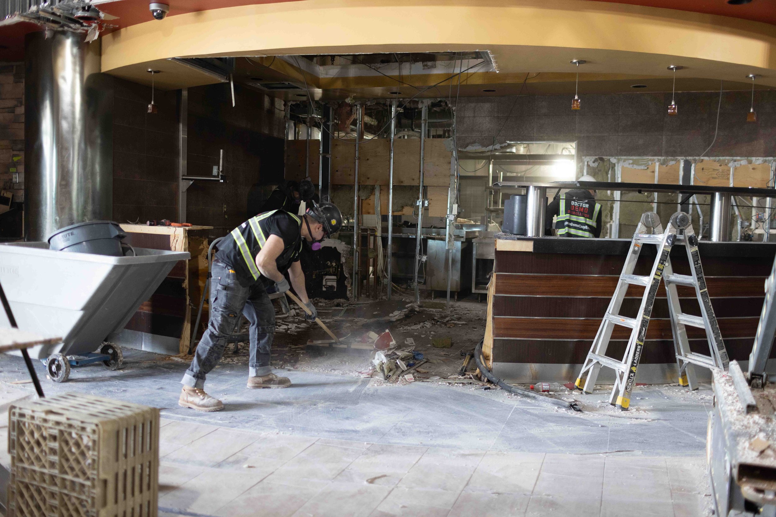 Workers demolishing the interior of a restaurant or store, with debris on the floor, partially dismantled walls, and exposed ceiling fixtures.