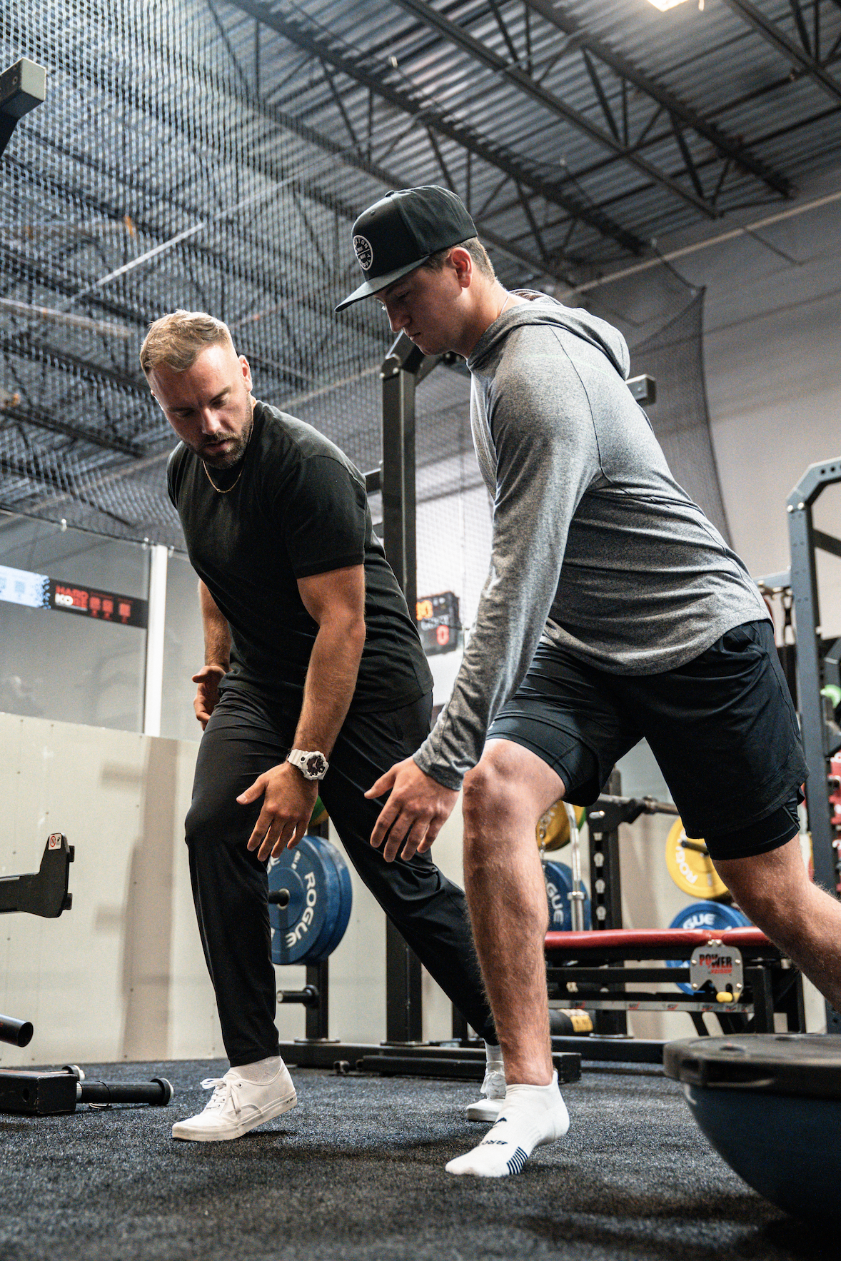 Two young men in workout attire performing a stretching exercise in a gym. One wears a gray hoodie, black shorts, and a baseball cap; the other wears a black t-shirt, black pants, and white sneakers. Gym equipment, weights, and a black floor mat are visible in the background.