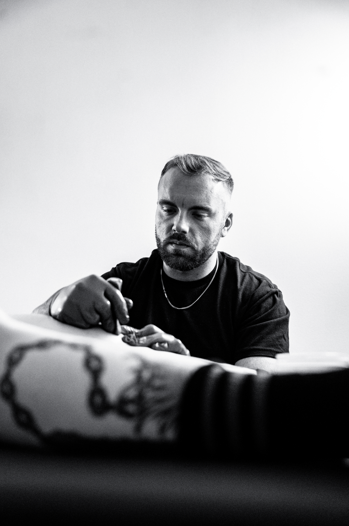 A black and white photo of a man with short hair and a beard, wearing a black t-shirt and gloves, focused on working on a tattoo machine.