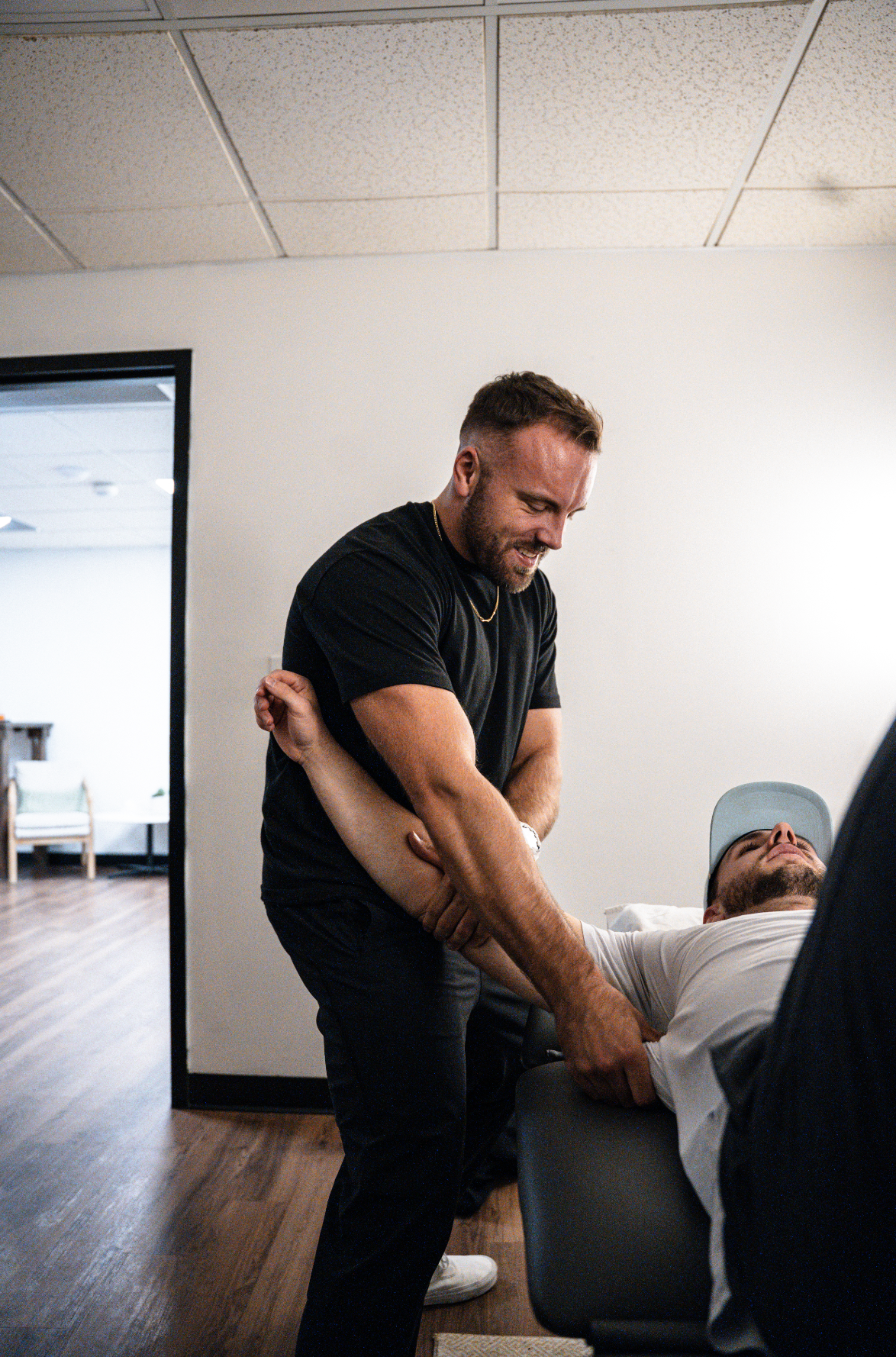 Two men engaged in a playful or lighthearted activity in an indoor setting. One man is standing and holding onto the other man, who is reclining in a chair, both smiling. The setting appears to be an office or medical environment with plain walls and ceiling tiles.