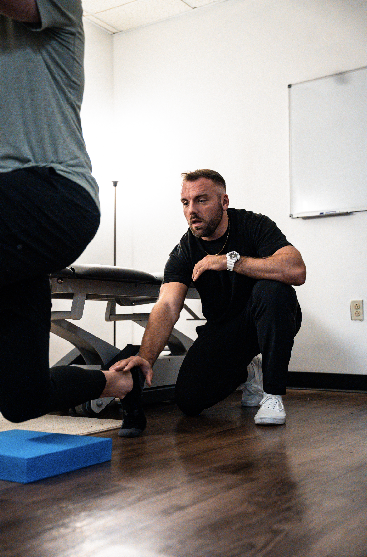 A man in black clothing and white sneakers is kneeling on the floor, holding a person's foot, and appears to be giving physical therapy or stretching treatment to someone sitting or lying on a massage table in a room with white walls and a whiteboard.