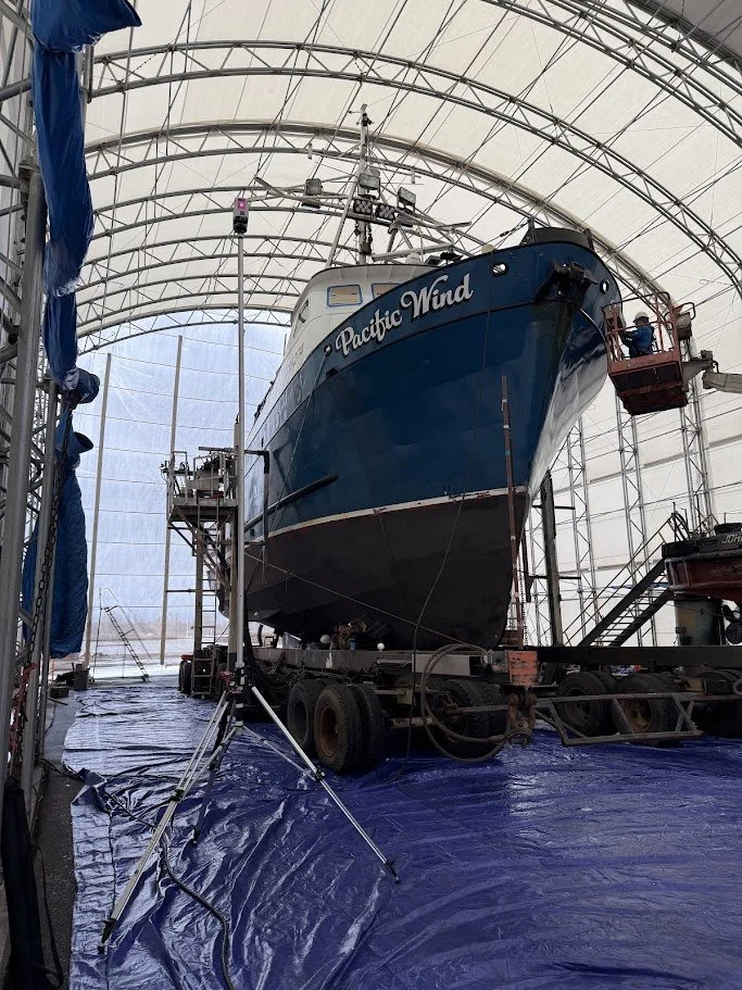 A large fishing boat named 'Pacific Wind' is inside a covered boatyard on a trailer, with scaffolding and maintenance equipment around it.