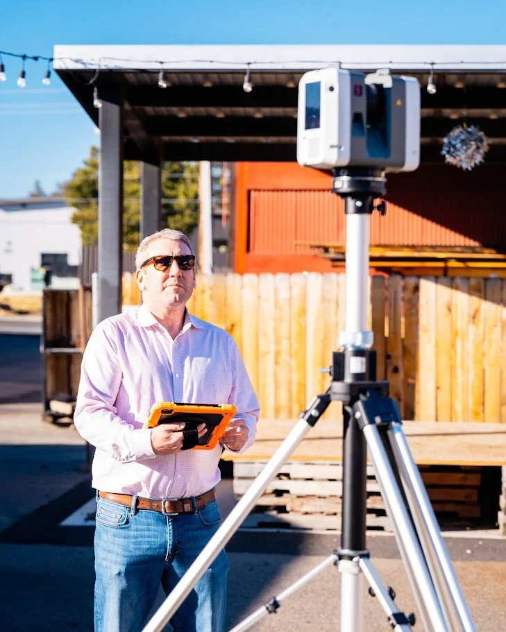 Man with sunglasses holding a device in front of a 3D laser scanner mounted on a tripod outdoors