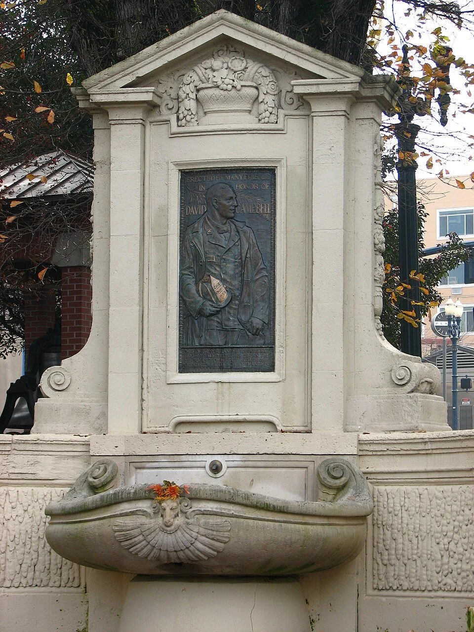 A monument with a memorial plaque and a fountain. The plaque features a relief of a man in military uniform, and the fountain has a decorative face with a flower decoration.