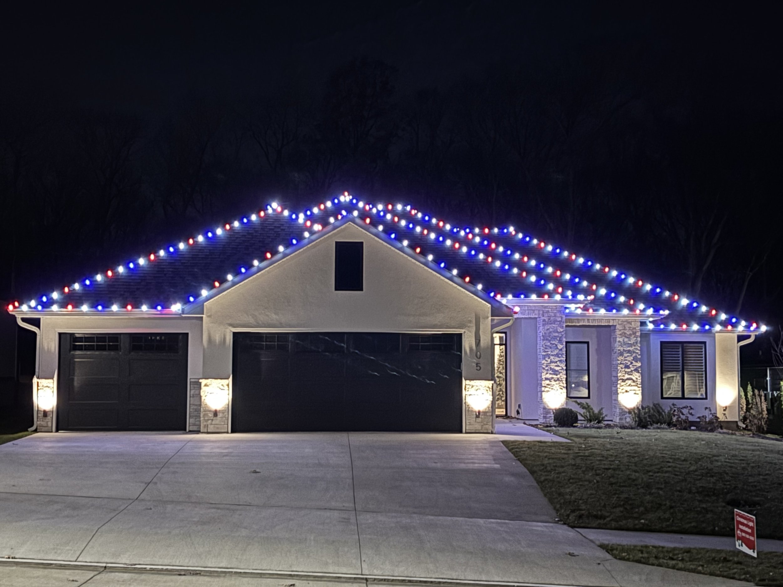 A modern house decorated with red, white, and blue string lights along the roof at night. The house has a two-car garage and a small front lawn.