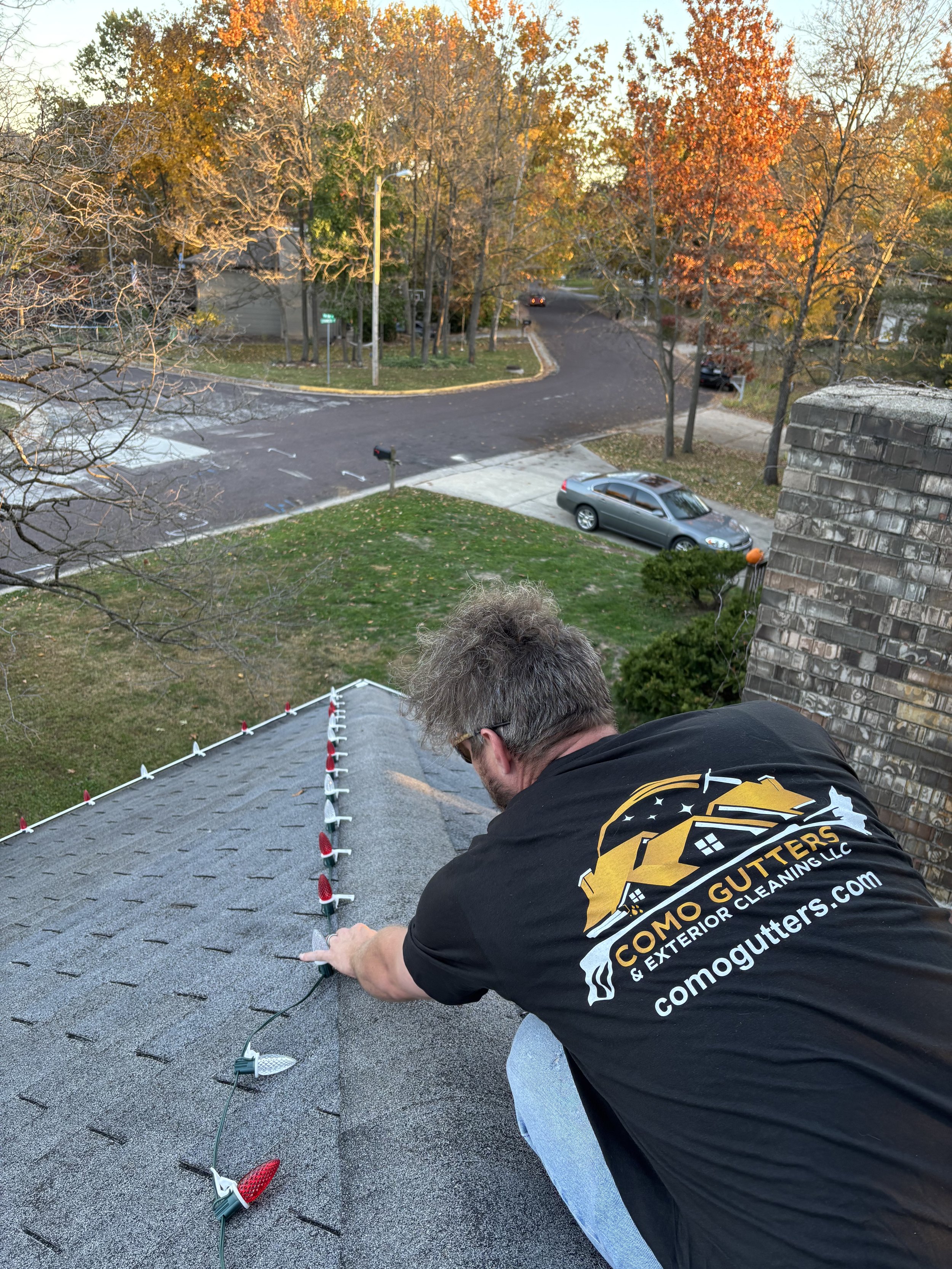 A man installing Christmas lights on a roof at sunset, wearing a black shirt with company branding, with a street, trees with autumn foliage, and parked cars in the background.