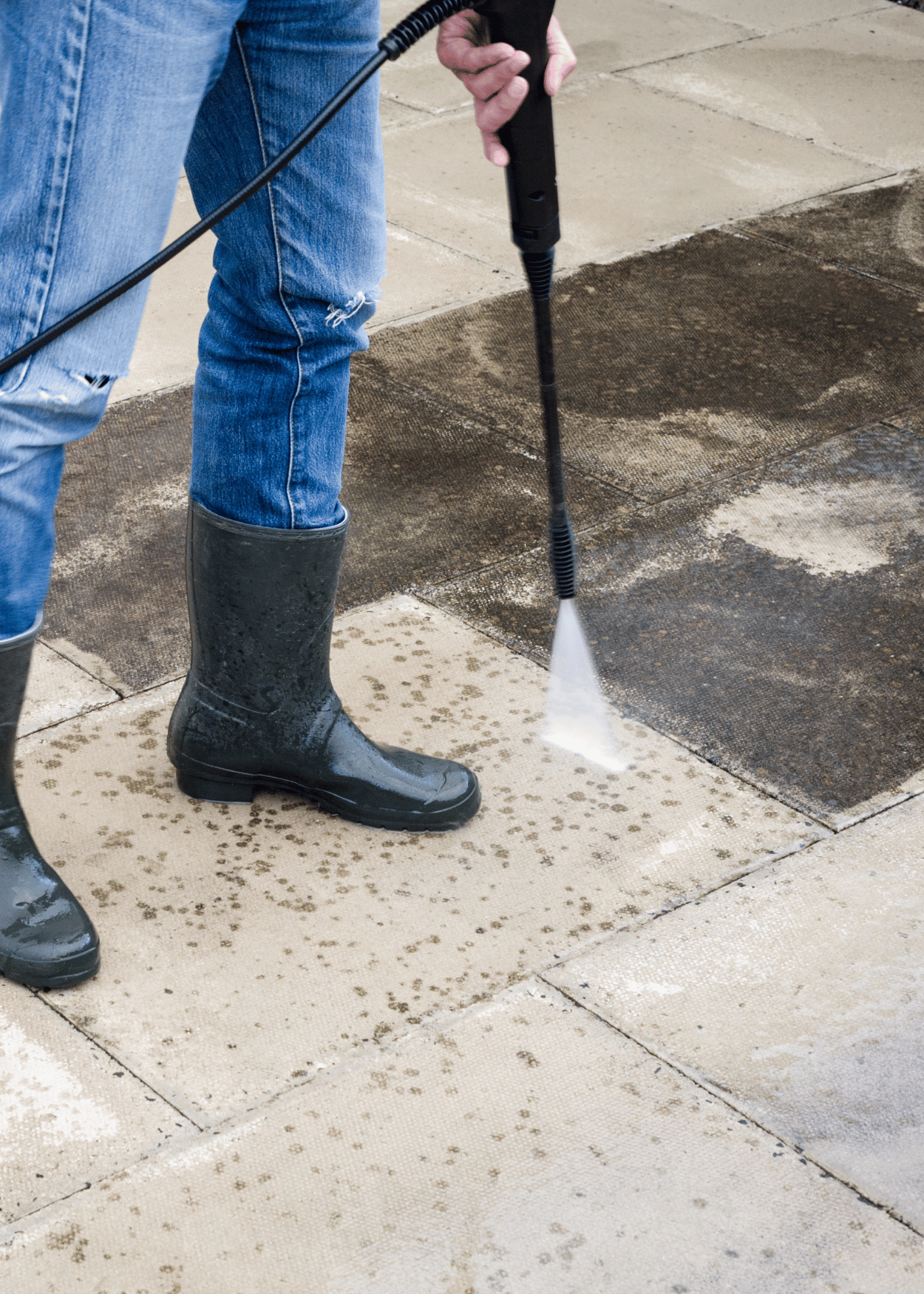 Person wearing black rubber boots and blue jeans pressure washing a concrete sidewalk.