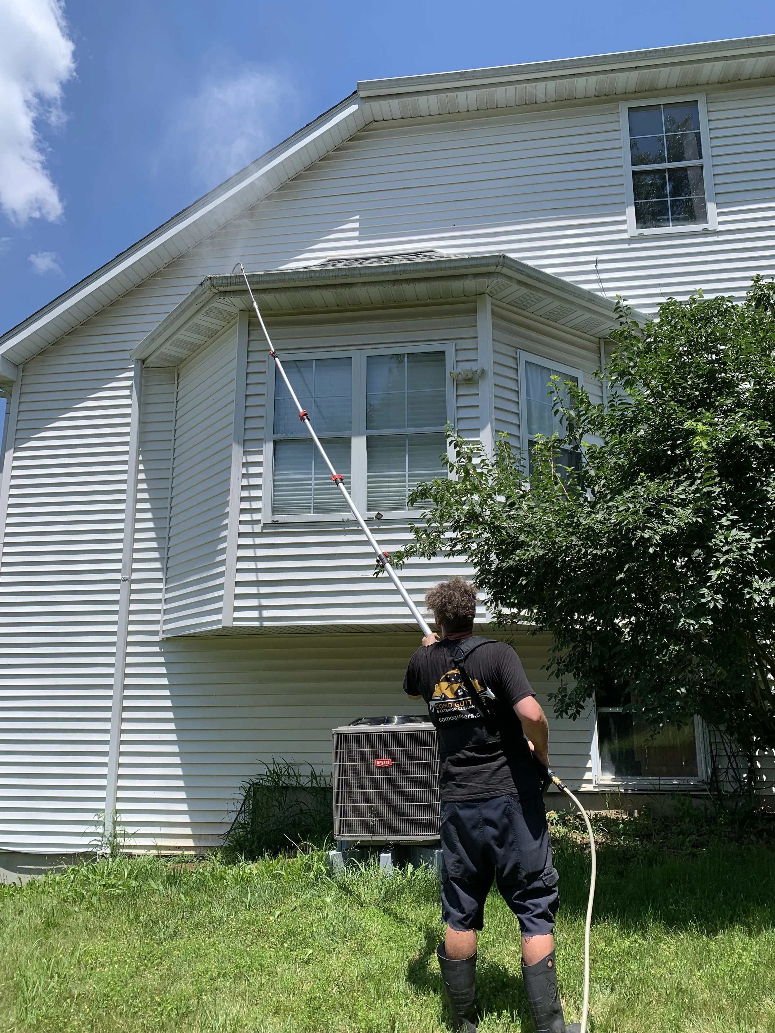 A person cleaning the exterior windows of a two-story house with a long pole, standing on a grassy lawn with a tree nearby and an air conditioning unit in the background under a blue sky with some clouds.