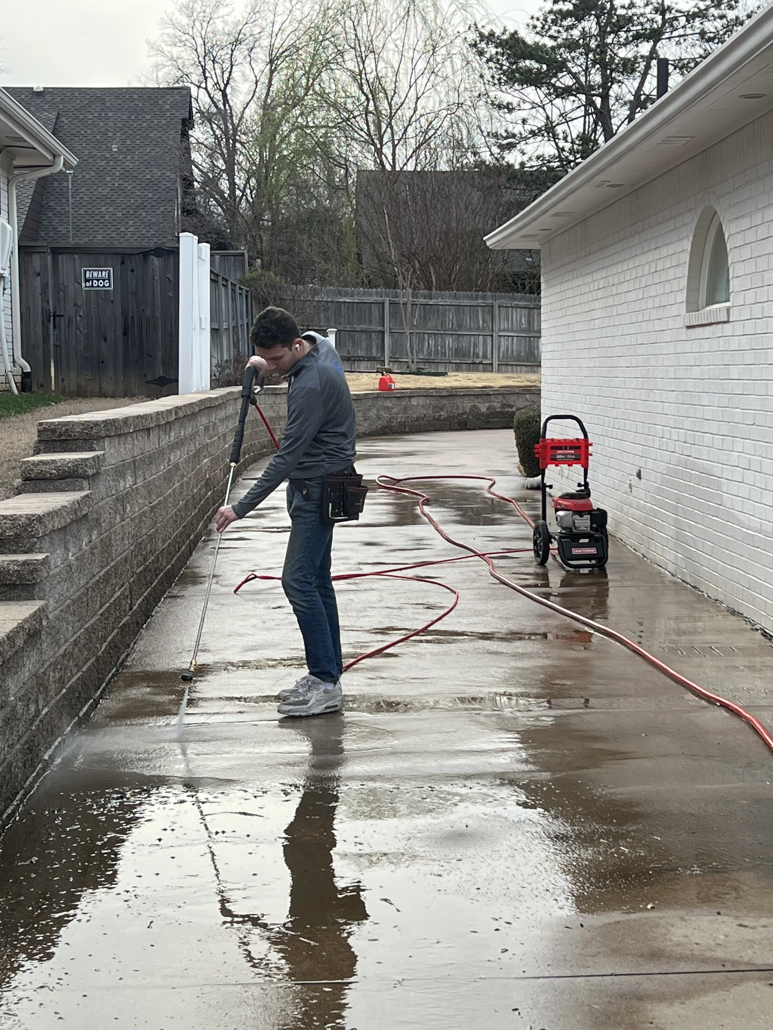 A person is pressure washing a concrete driveway in a backyard. The driveway has water and soap on it, and there is equipment including a pressure washer on a cart nearby. The background shows a wooden fence, a white house, and some trees.