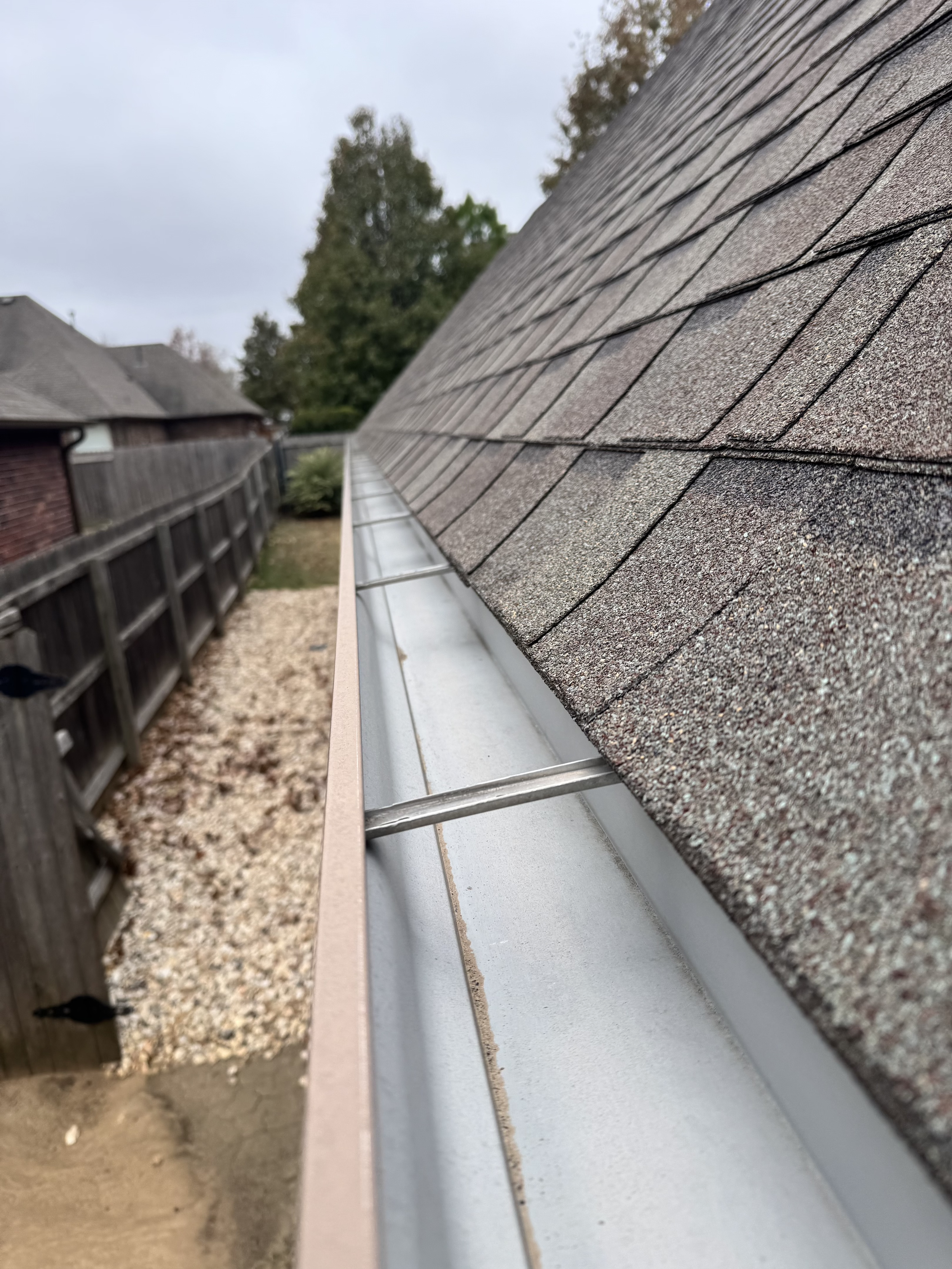 Close-up of a roof with asphalt shingles, a metal flashing installed along the edge, and a gutter underneath, with a wooden fence and neighborhood houses in the background.