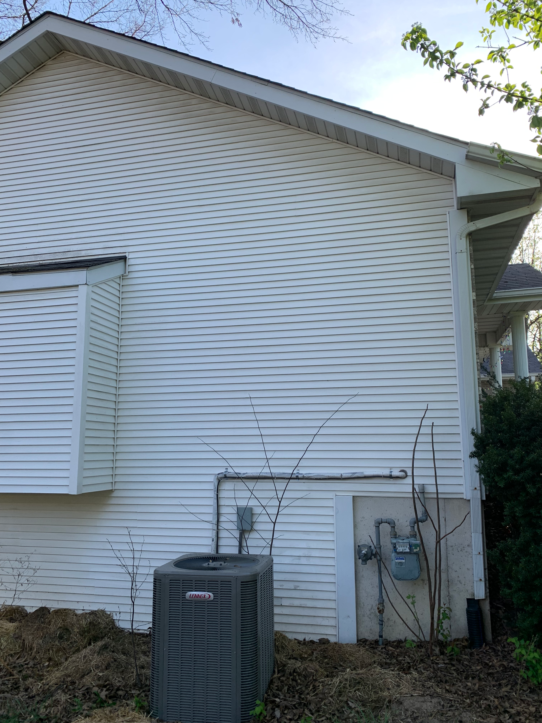 Back side of a house showing beige vinyl siding, a downspout, an air conditioning unit, a utility meter, and some small trees and bushes.