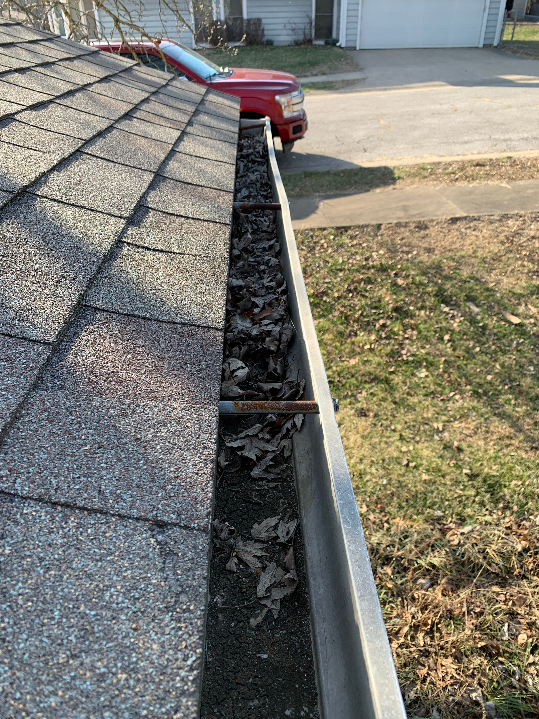 View of a roof's gutter filled with dried leaves, seen from the shingles' edge with a parked red truck and a house with a garage in the background.
