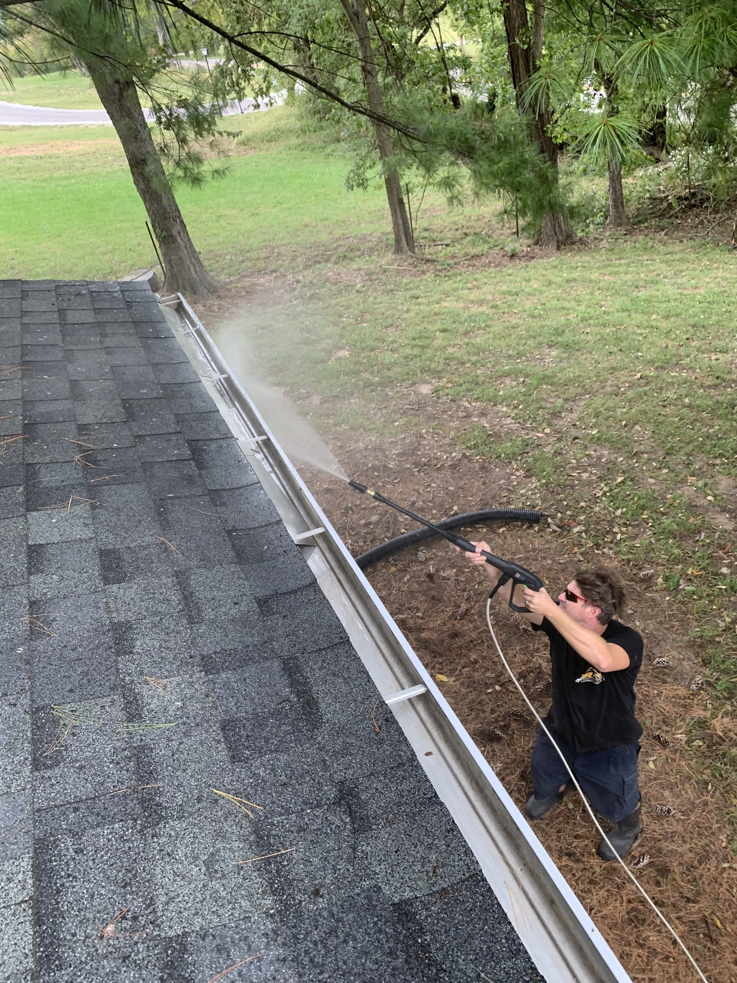 A man wearing sunglasses and a black t-shirt is pressure washing the roof of a house. The house has gray shingles, and the man is holding a long pressure washer wand with water coming out. There are trees and greenery in the background.