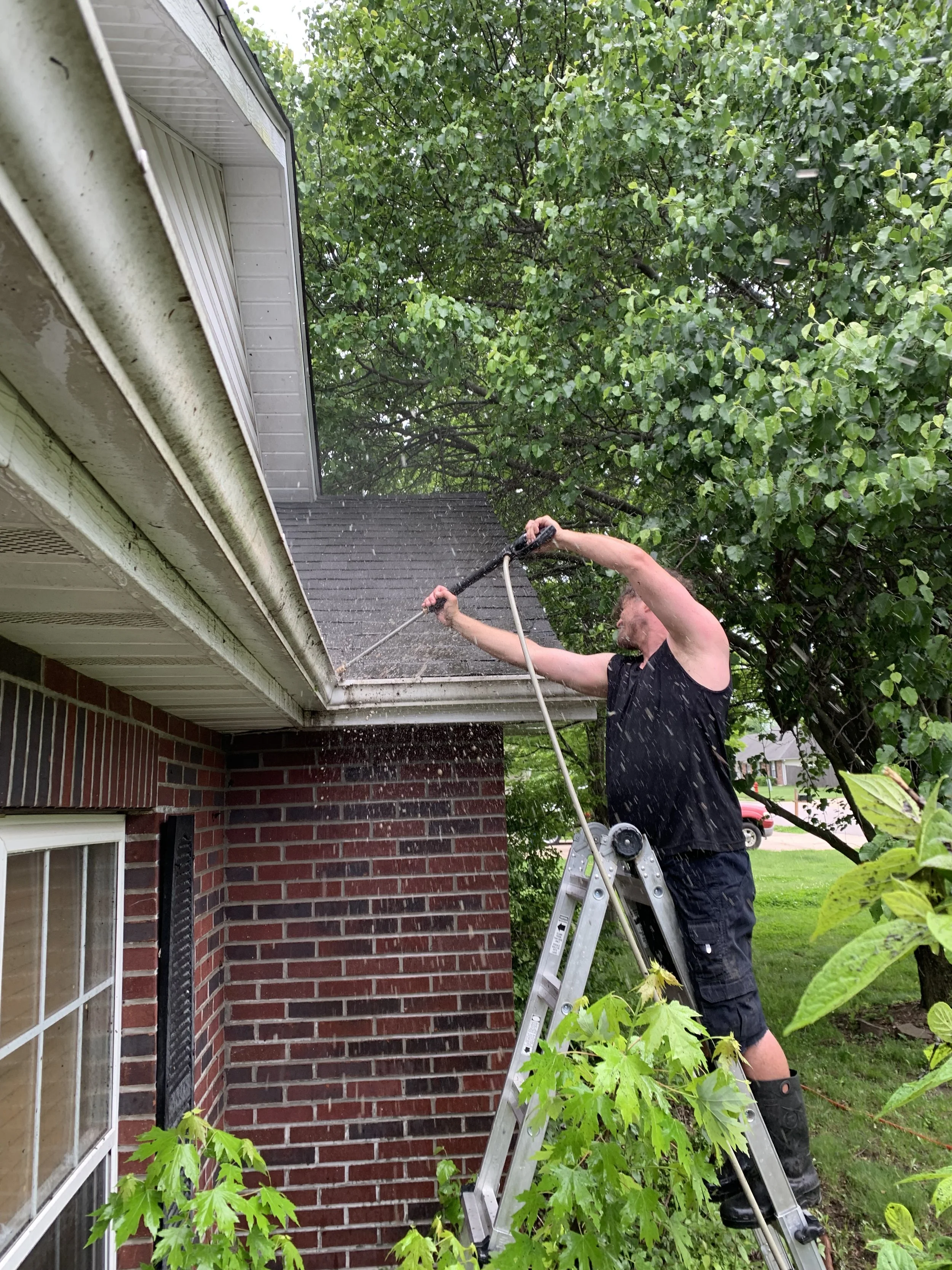 A man standing on a ladder pressure washing the roof of a house with a garden and trees in the background.